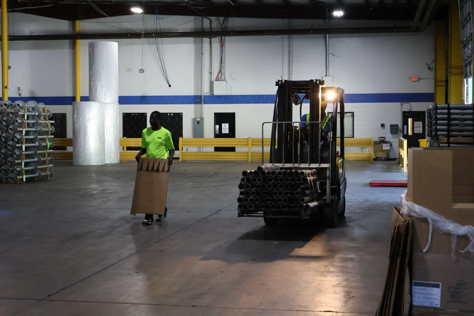 Two workers in a warehouse, one walking while carrying cardboard, the other operating a forklift with metal pipes, surrounded by stacked materials and boxes.
