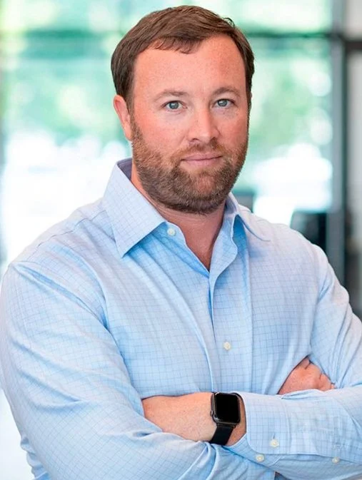 Man with short beard and light blue shirt standing with arms crossed, wearing a smartwatch.