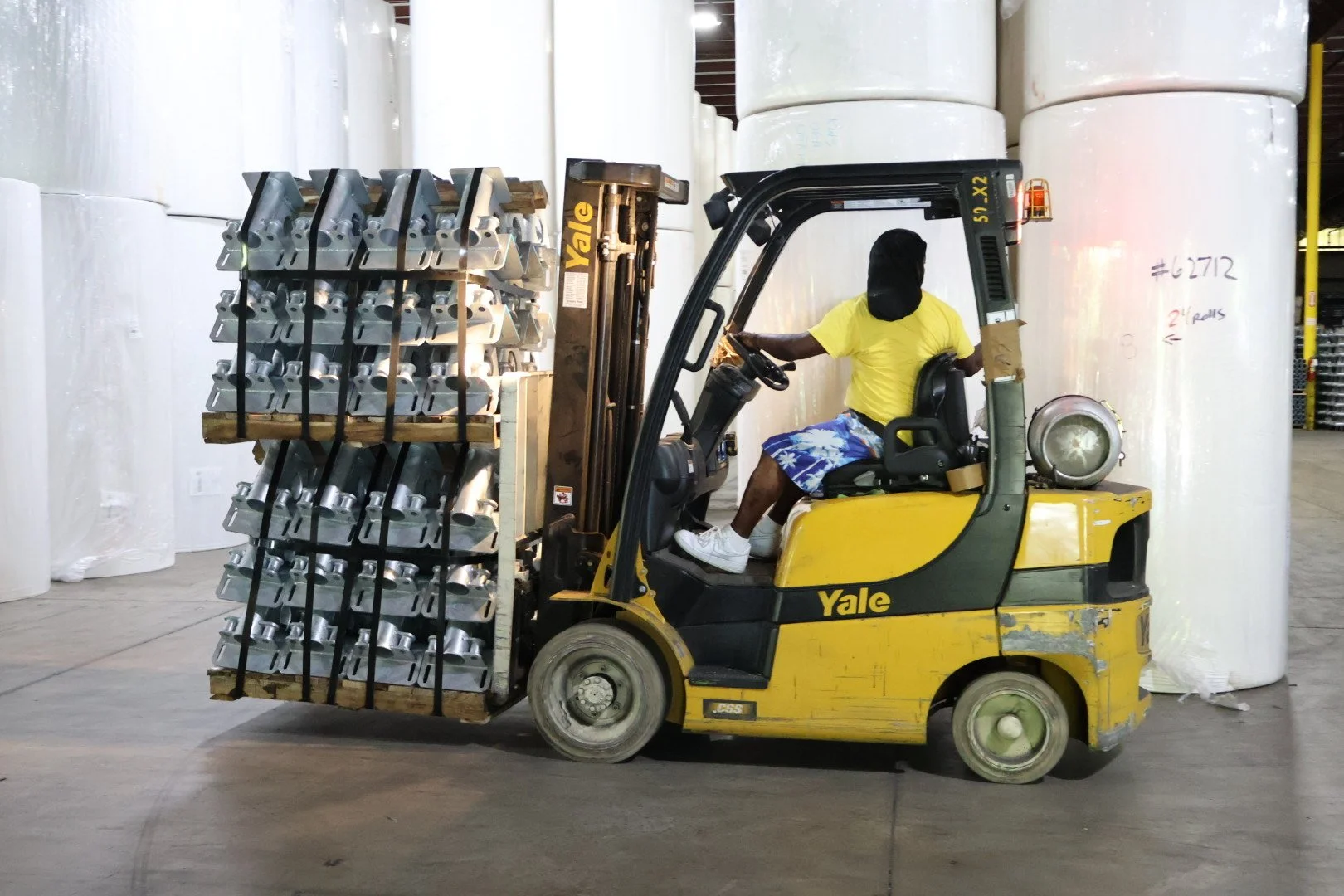 A person driving a yellow Yale forklift carrying a pallet of metal pieces inside a warehouse with large rolls of plastic or fabric in the background.