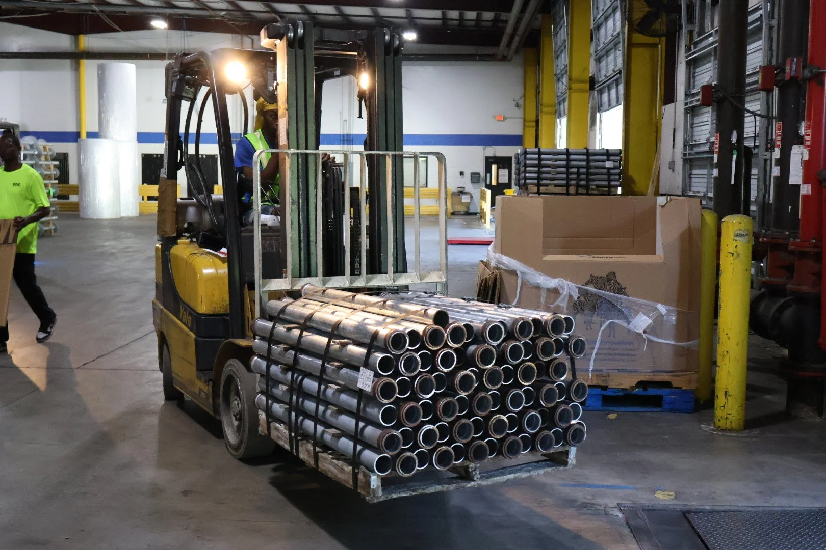 A forklift carrying metal pipes in a warehouse or industrial setting with workers and storage areas visible in the background.