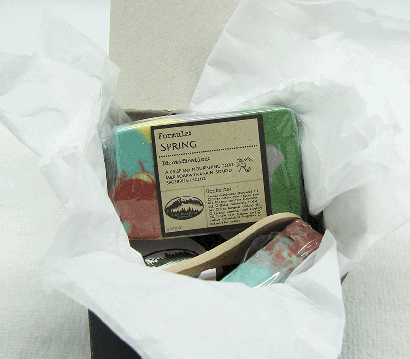 Spring Collection box set nestled in white tissue paper inside a black gift box showing wrapped soap bars, wooden cosmetic spoon, and other products packed together