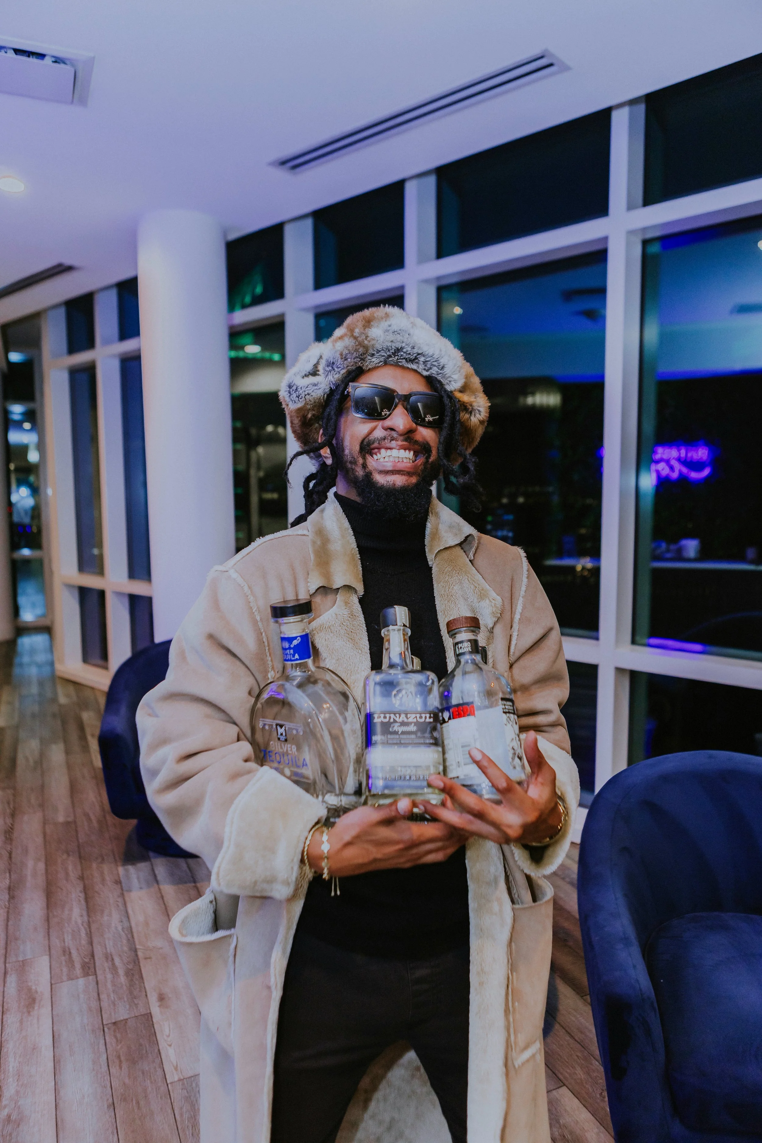 A man wearing sunglasses, a fur hat, and a beige coat is smiling and holding multiple bottles of alcohol in a room with large windows and modern decor.