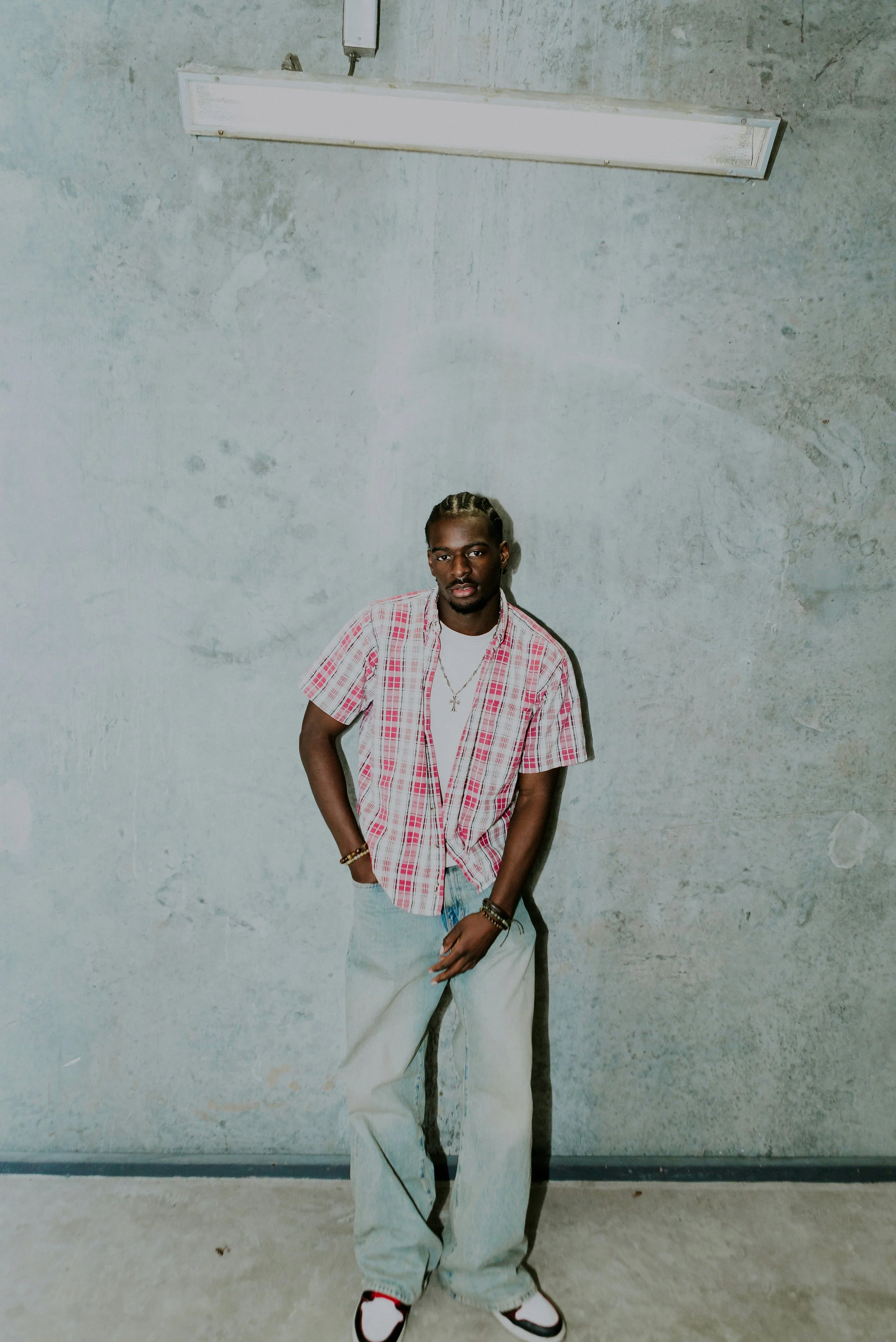 A young man with dark skin and braided hair, wearing a red and white plaid short-sleeve shirt over a white T-shirt, light-colored baggy jeans, and white sneakers with red accents, standing against a plain concrete wall.