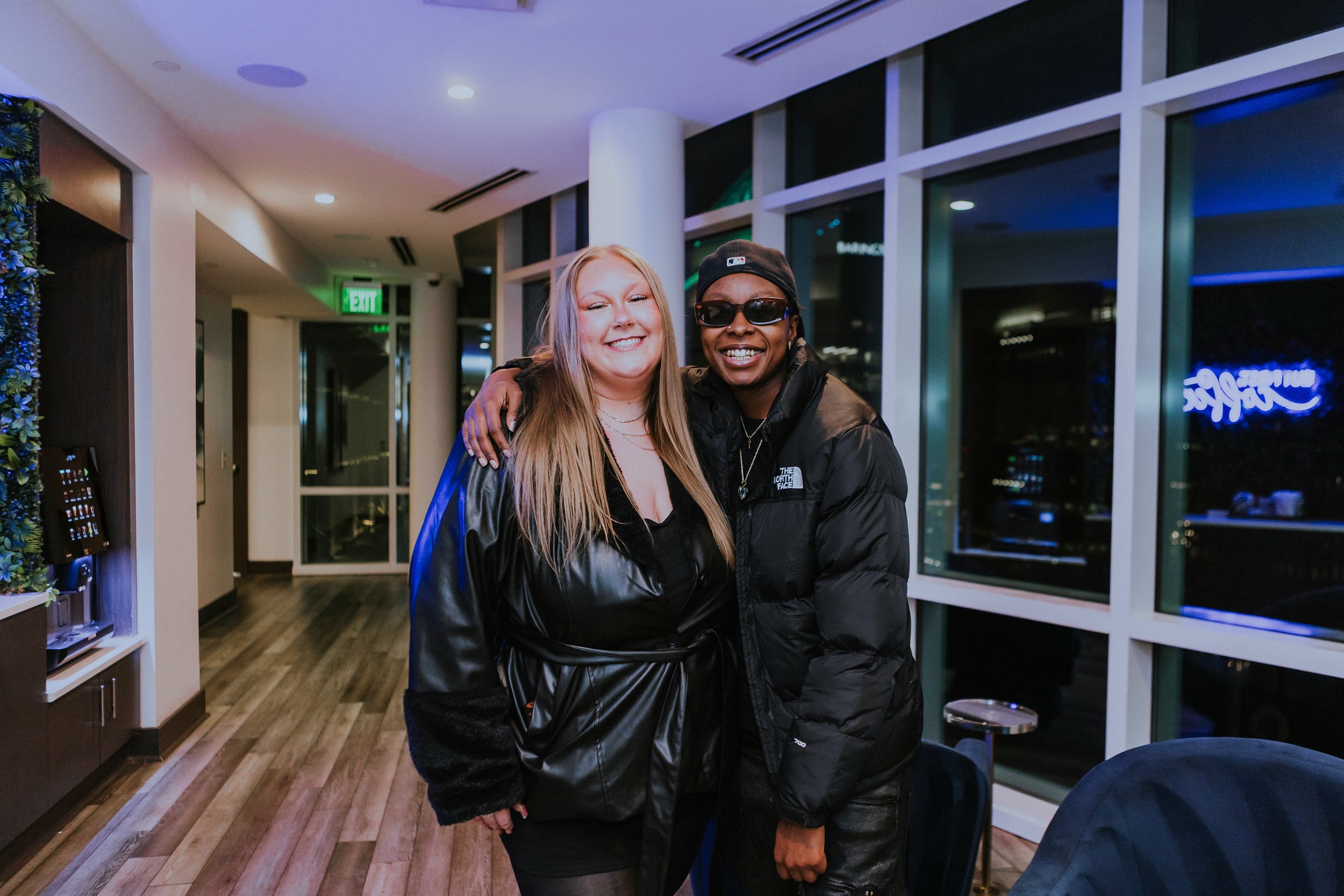 Two people smiling and posing for a photo inside a modern building at night. One person has long blonde hair and the other is wearing sunglasses and a black cap.