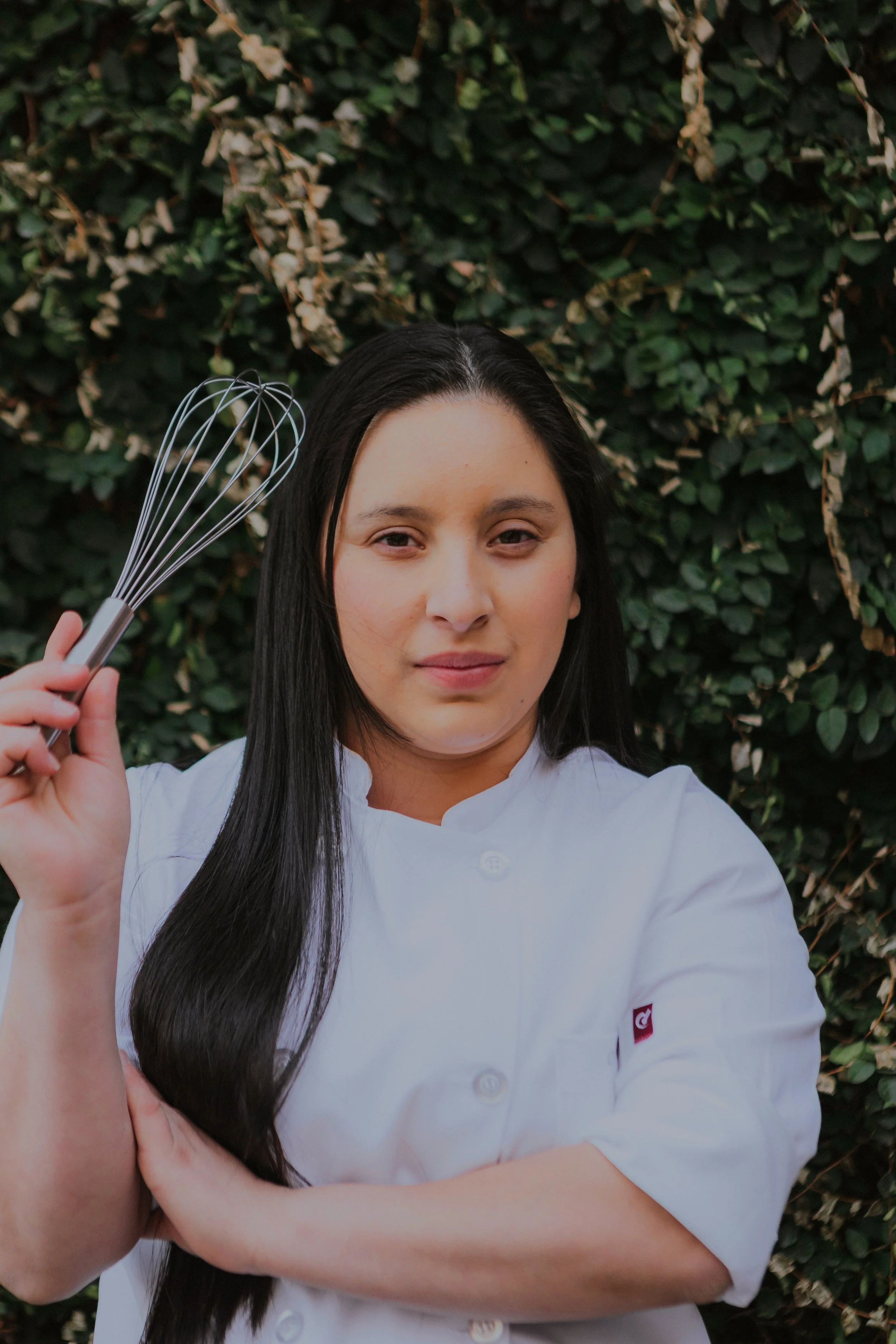 A female chef with long dark hair, wearing a white chef's coat, holding a whisk in her right hand while standing in front of a lush green leafy background.