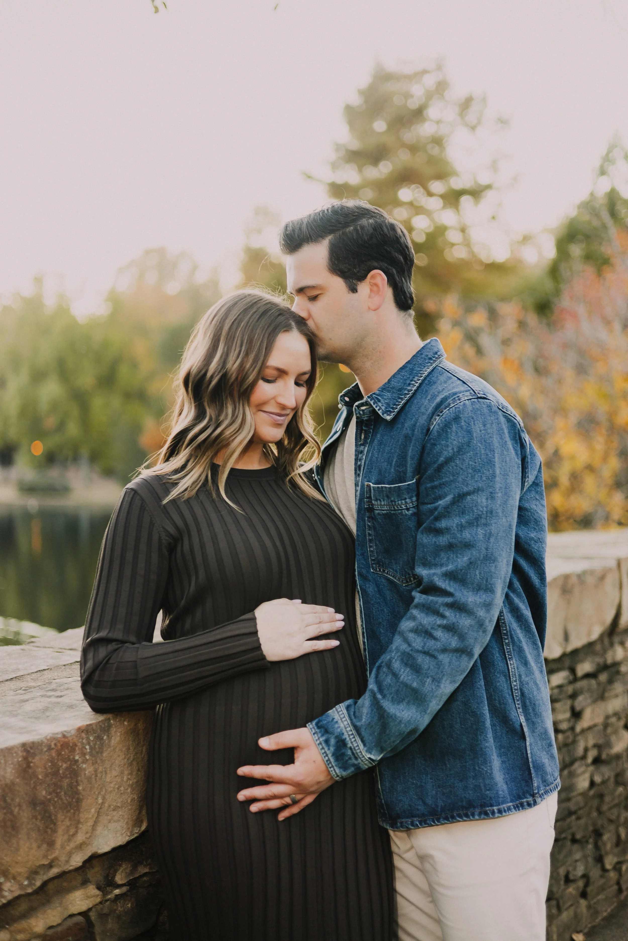 A pregnant woman and a man are standing close together by a stone wall near a body of water, with trees in the background. The woman has her hand on her belly, and the man is gently holding her belly, while he kisses her forehead.