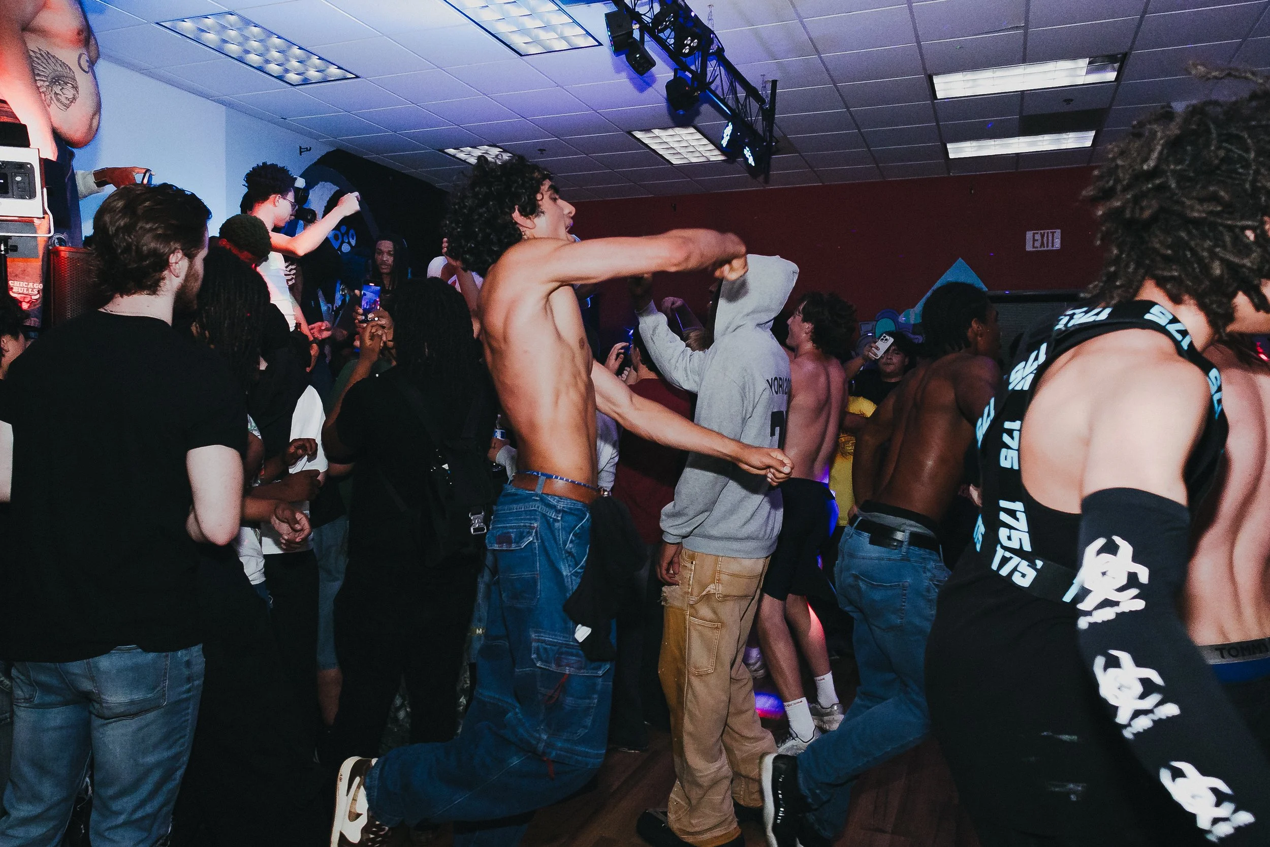 People dancing and having fun at a crowded indoor concert or club, with some shirtless men and others taking photos.