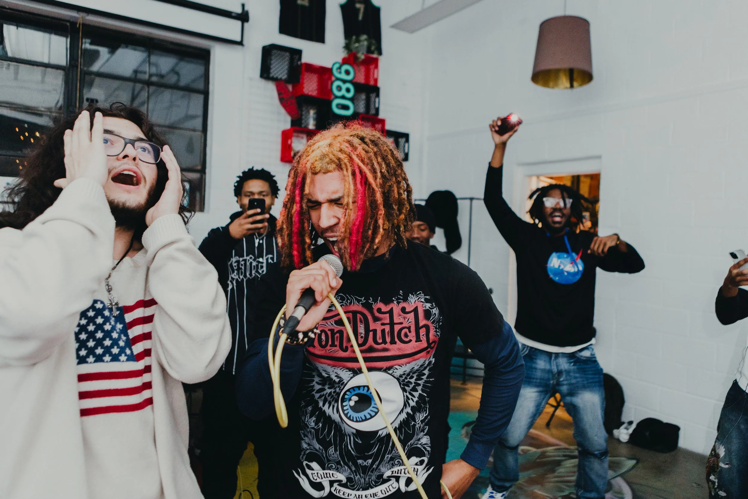 A group of young people enjoying lively music and singing at an indoor party or gathering. One person with curly hair and glasses reacts with surprise, another with colorful dreadlocks passionately sings into a microphone, while others in the background are dancing or using their phones.