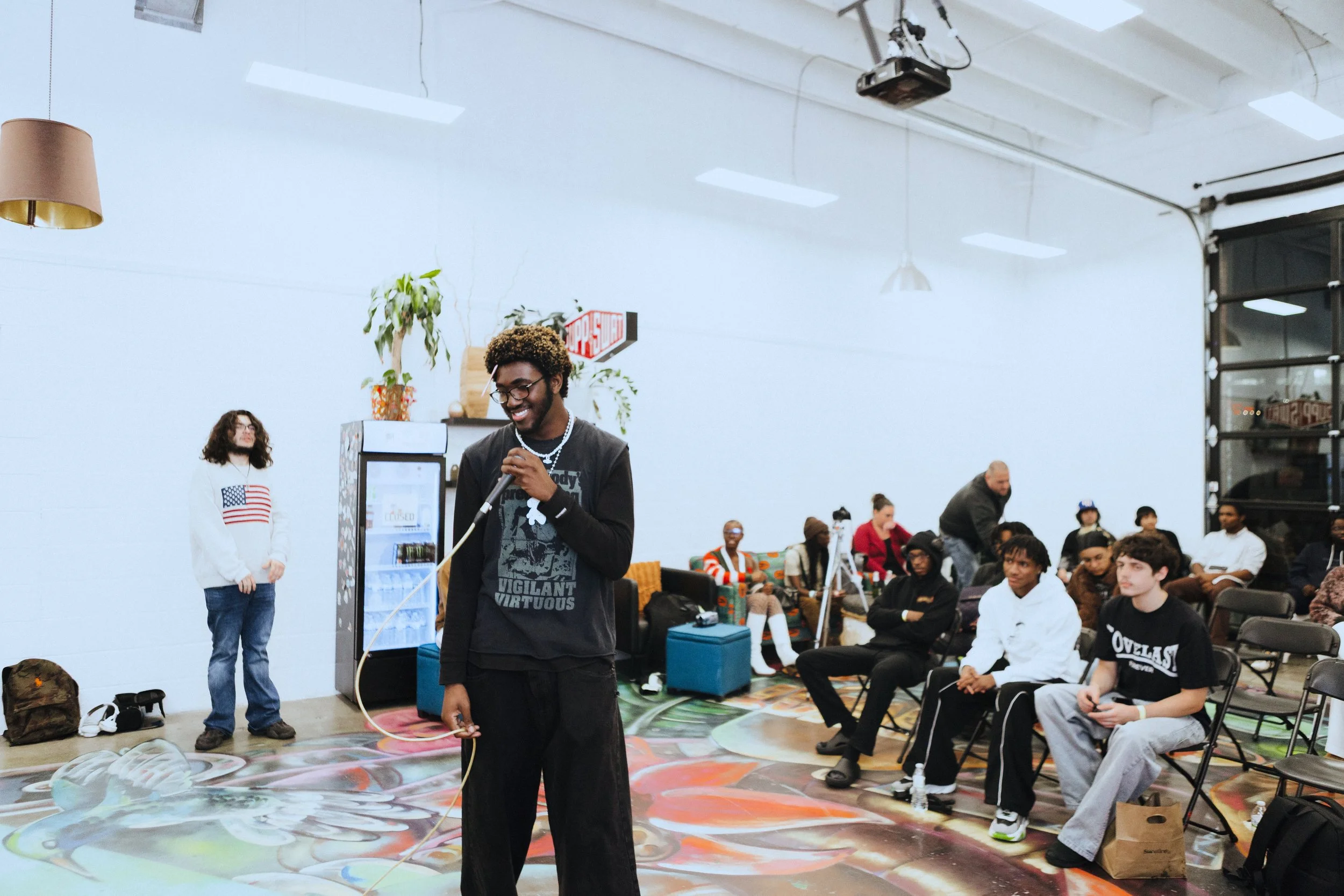 A group of people in a brightly lit indoor space, with a man holding a microphone on a colorful floor, and several seated audience members watching.