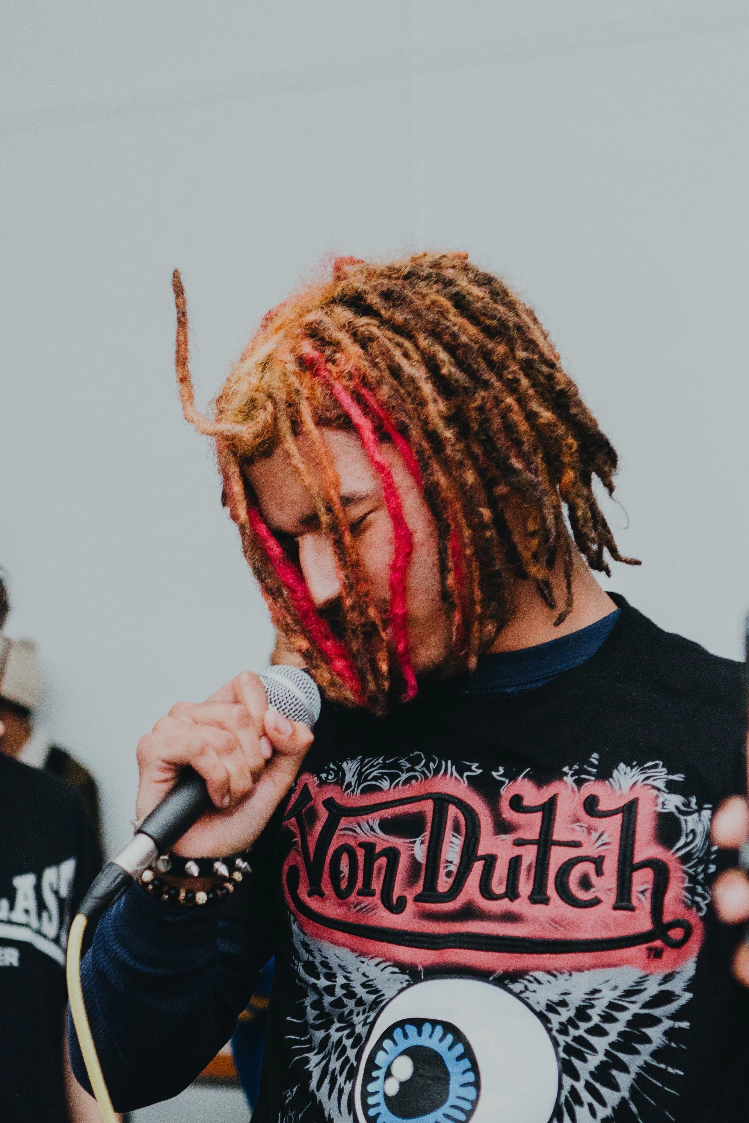 A person with dreadlocks dyed in red, brown, and blonde holding a microphone and singing or speaking. They are wearing a black shirt with a graphic design and the words "NonDutch" on it. There are other people in the background.