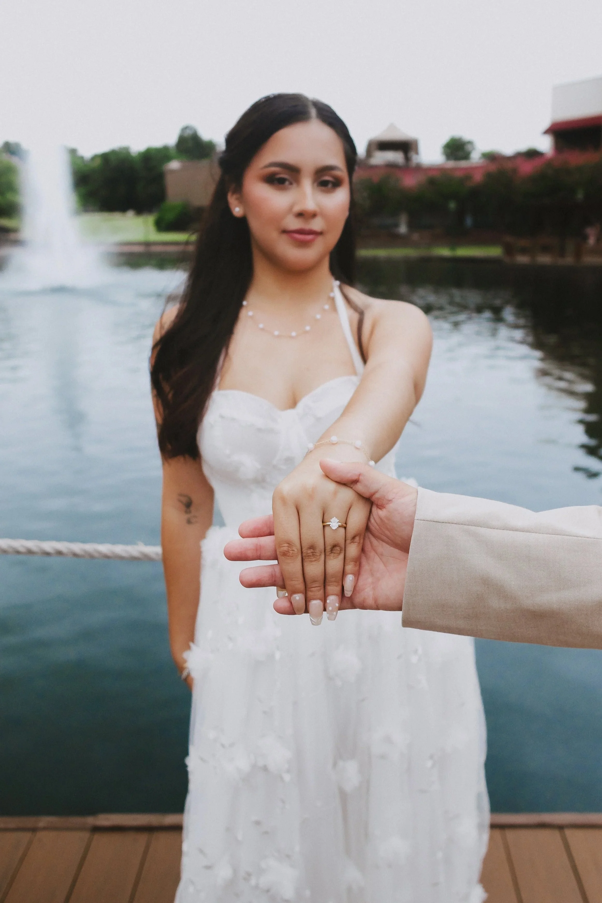 A woman in a white wedding dress showing a ring on her finger, with a man extending his hand to hold hers, standing by a lake or pond with trees and buildings in the background.