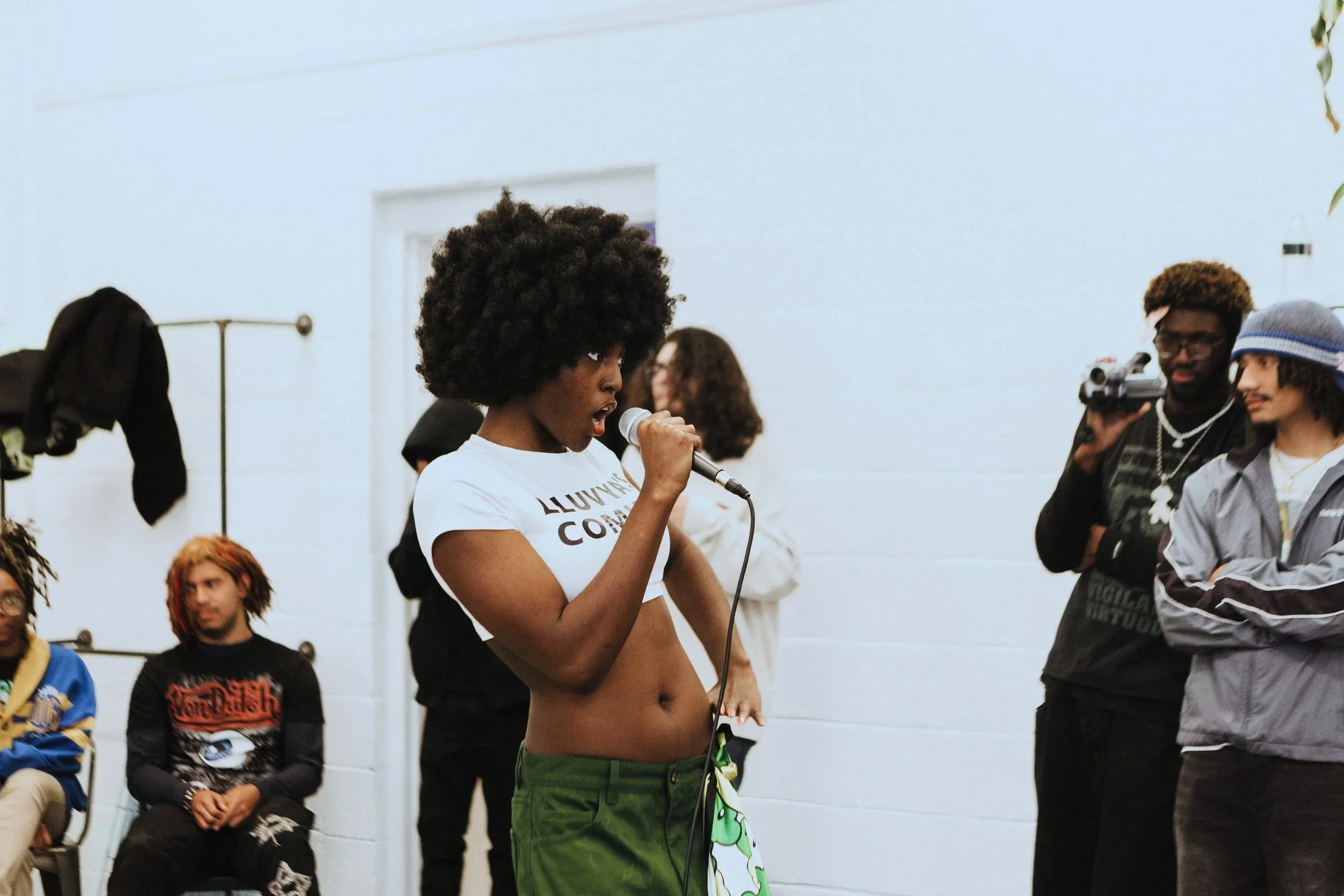 A young woman speaking into a microphone at an indoor gathering, with several people around her, some sitting and some standing, in a room with white walls.