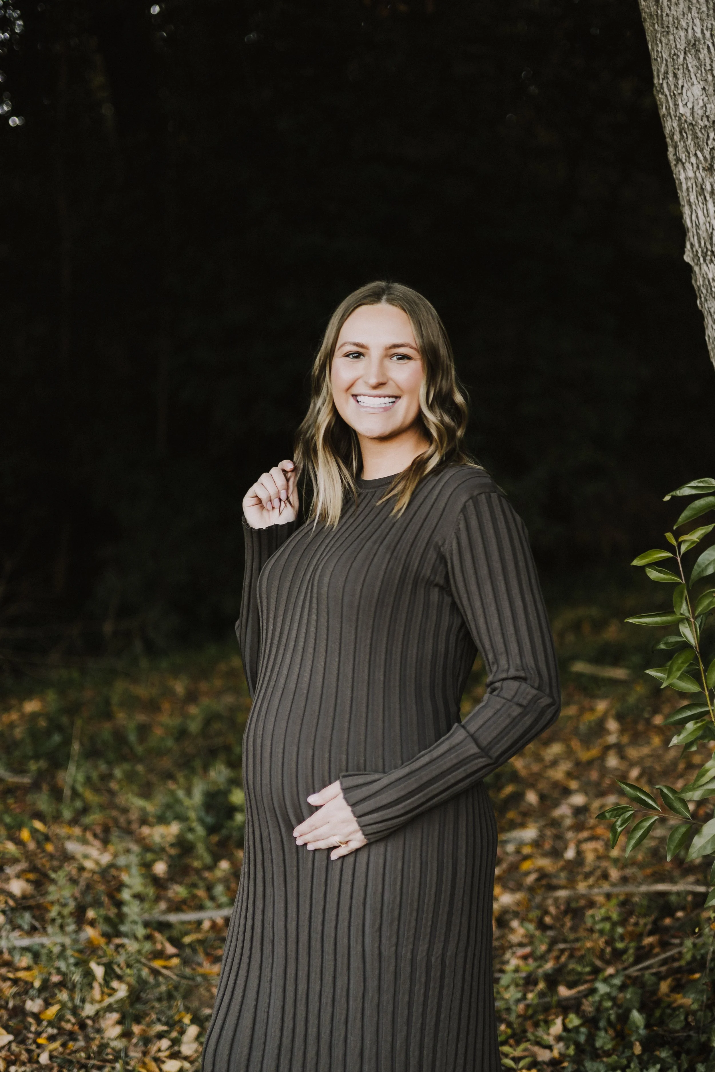 A smiling woman with shoulder-length wavy hair, wearing a long-sleeve, ribbed, brown dress, stands outdoors near a tree in a wooded area with fallen leaves on the ground.