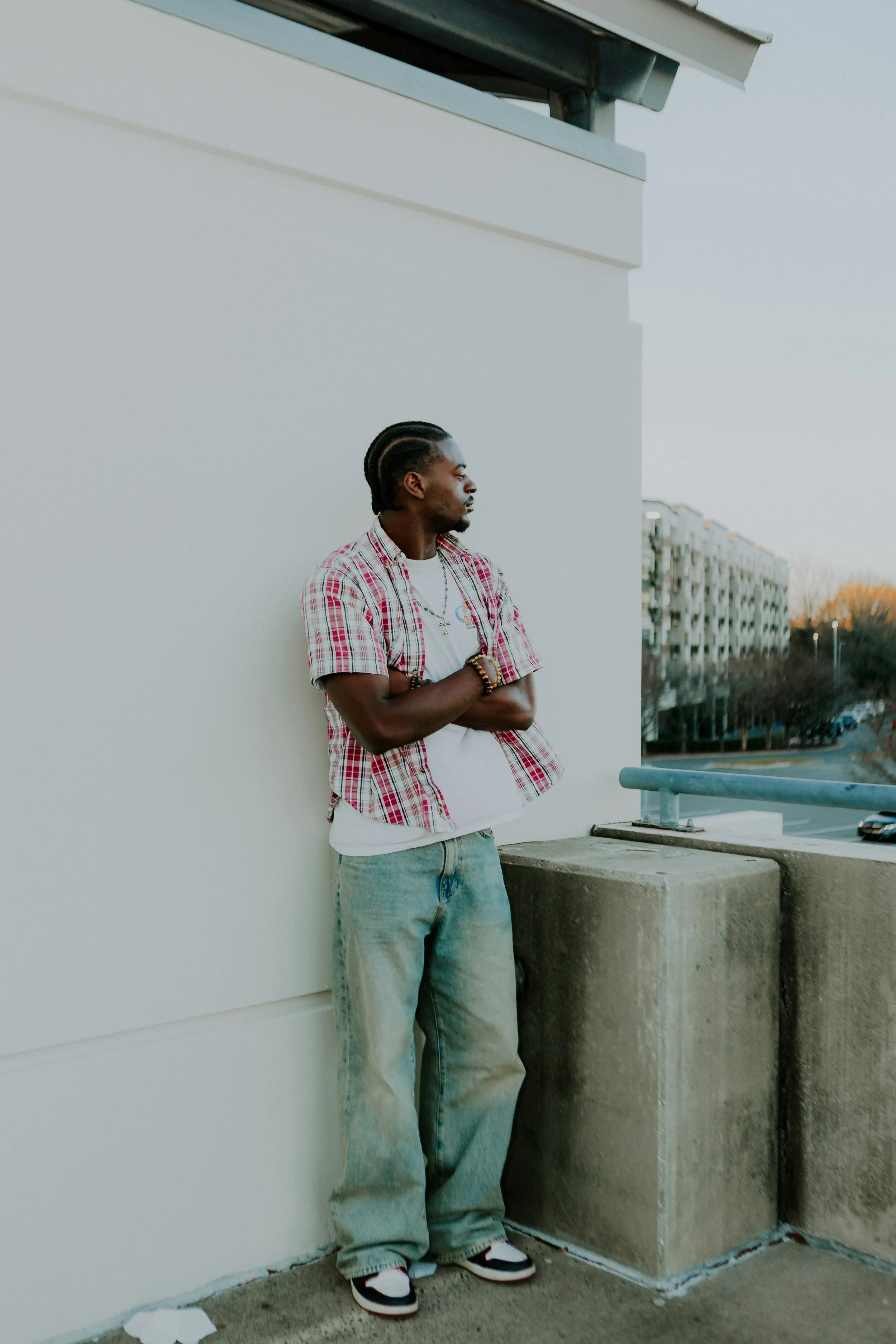 A young man standing on a rooftop balcony with arms crossed, looking to the side, wearing a red plaid shirt, white t-shirt, baggy jeans, and sneakers, against a cityscape background.