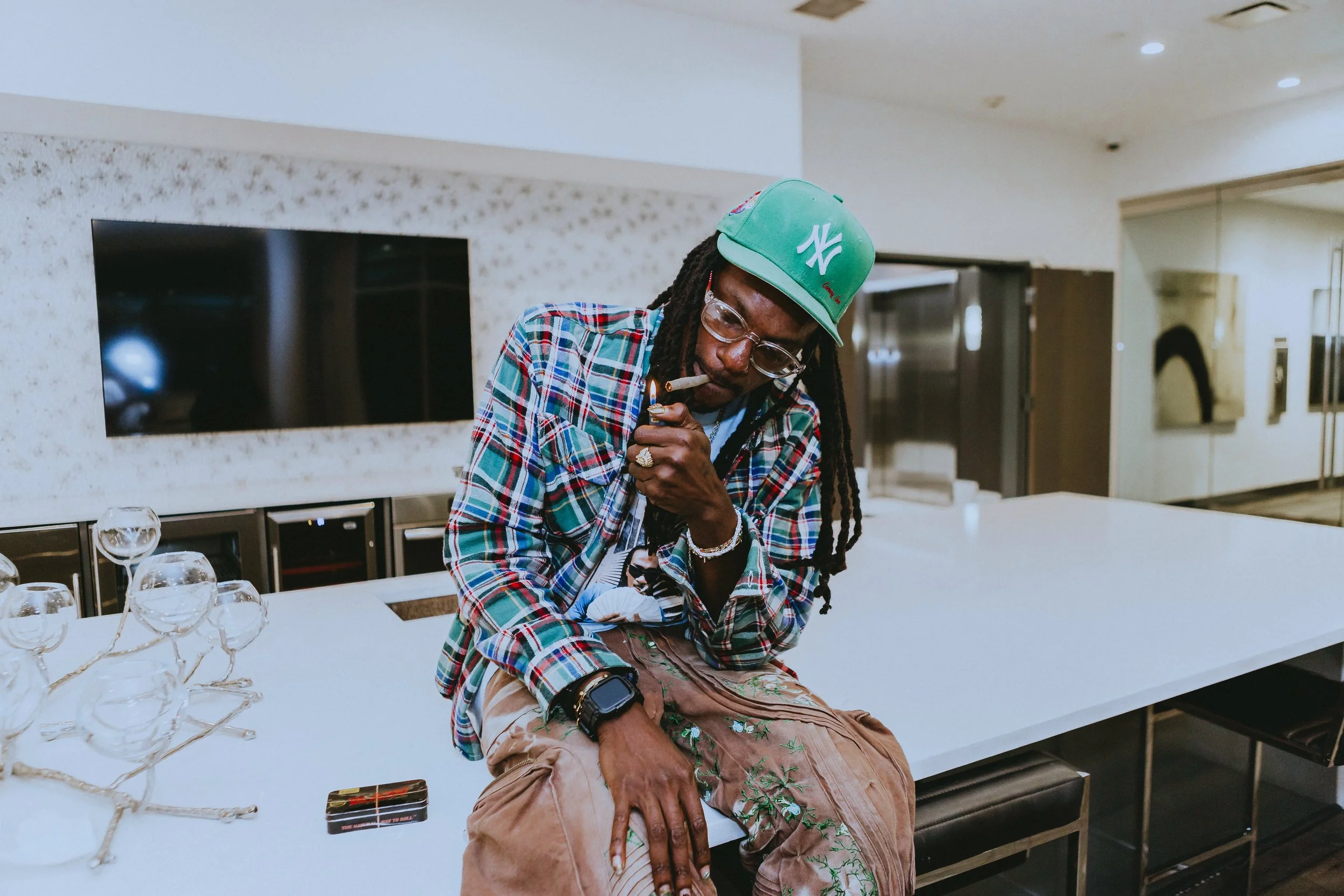 A man with dreadlocks, glasses, and jewelry is sitting on a table, lighting a cigarette, wearing a green New York Yankees cap, a plaid shirt, and beige embroidered pants, in a modern room with a white table and glassware.