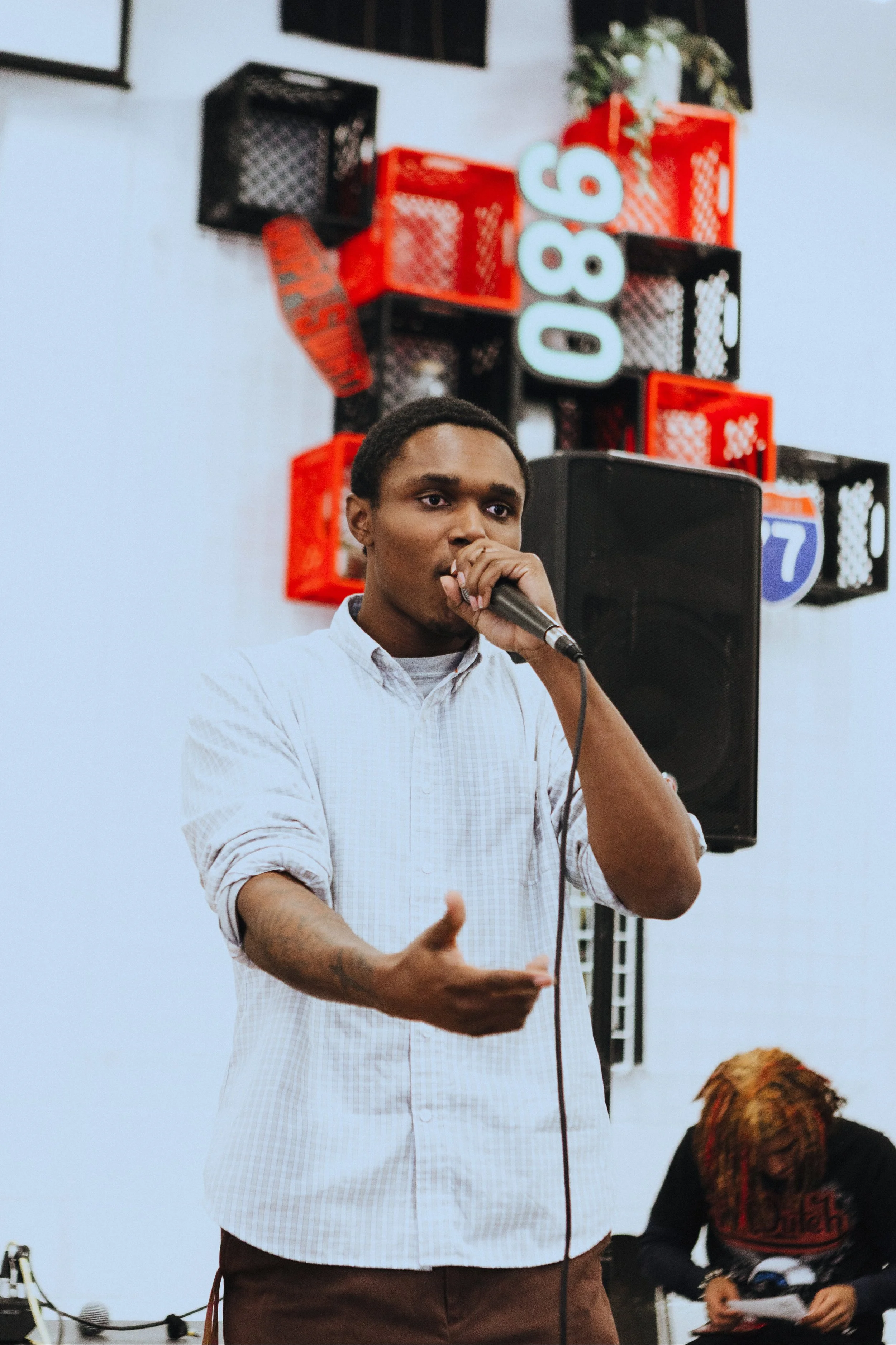 A young man in a white shirt speaking into a microphone at an indoor event, with a woman in the background reading from a paper. Red and black crates with neon signs in the shape of street signs are on the wall behind him.