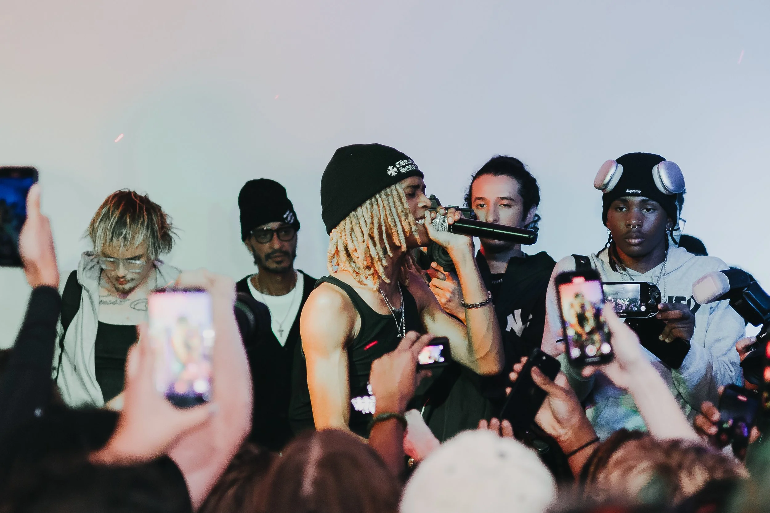 A woman with blond dreadlocks, wearing a black beanie and black tank top, performs with a microphone while surrounded by a crowd taking photos at an indoor event.