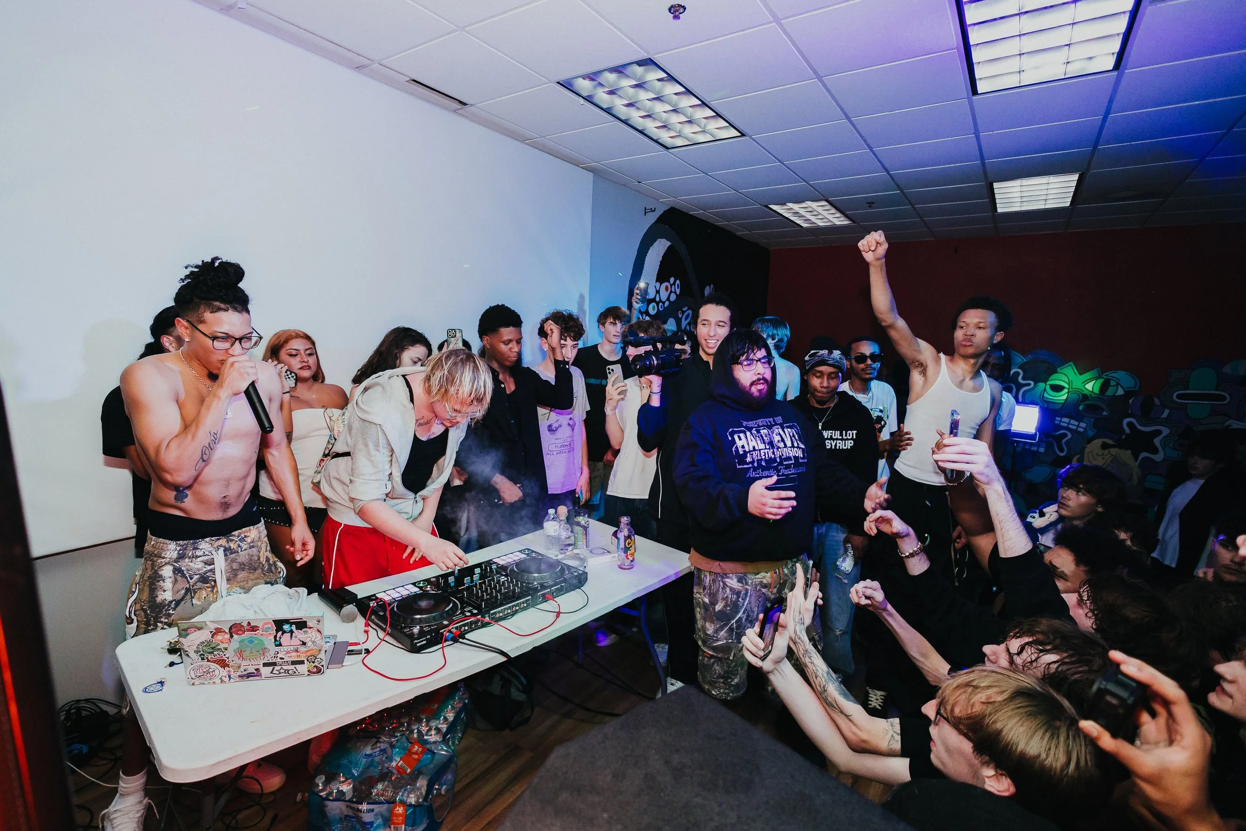 People dancing and socializing at a party with a DJ playing music on a mixer, some holding drinks, in a dimly lit room with colorful murals on the wall.