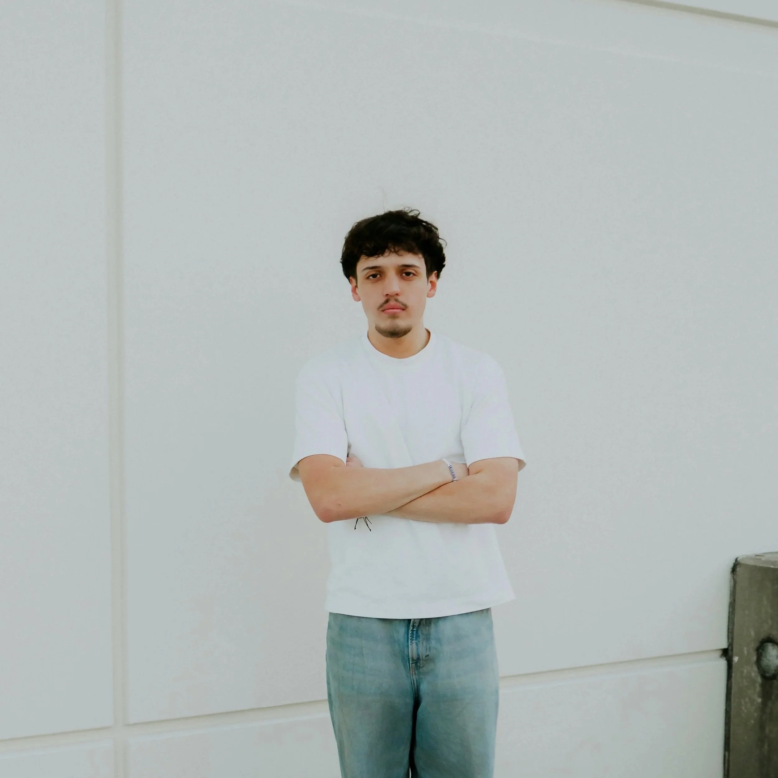 Young man standing against a blank white wall with crossed arms, wearing a white t-shirt and light blue jeans.