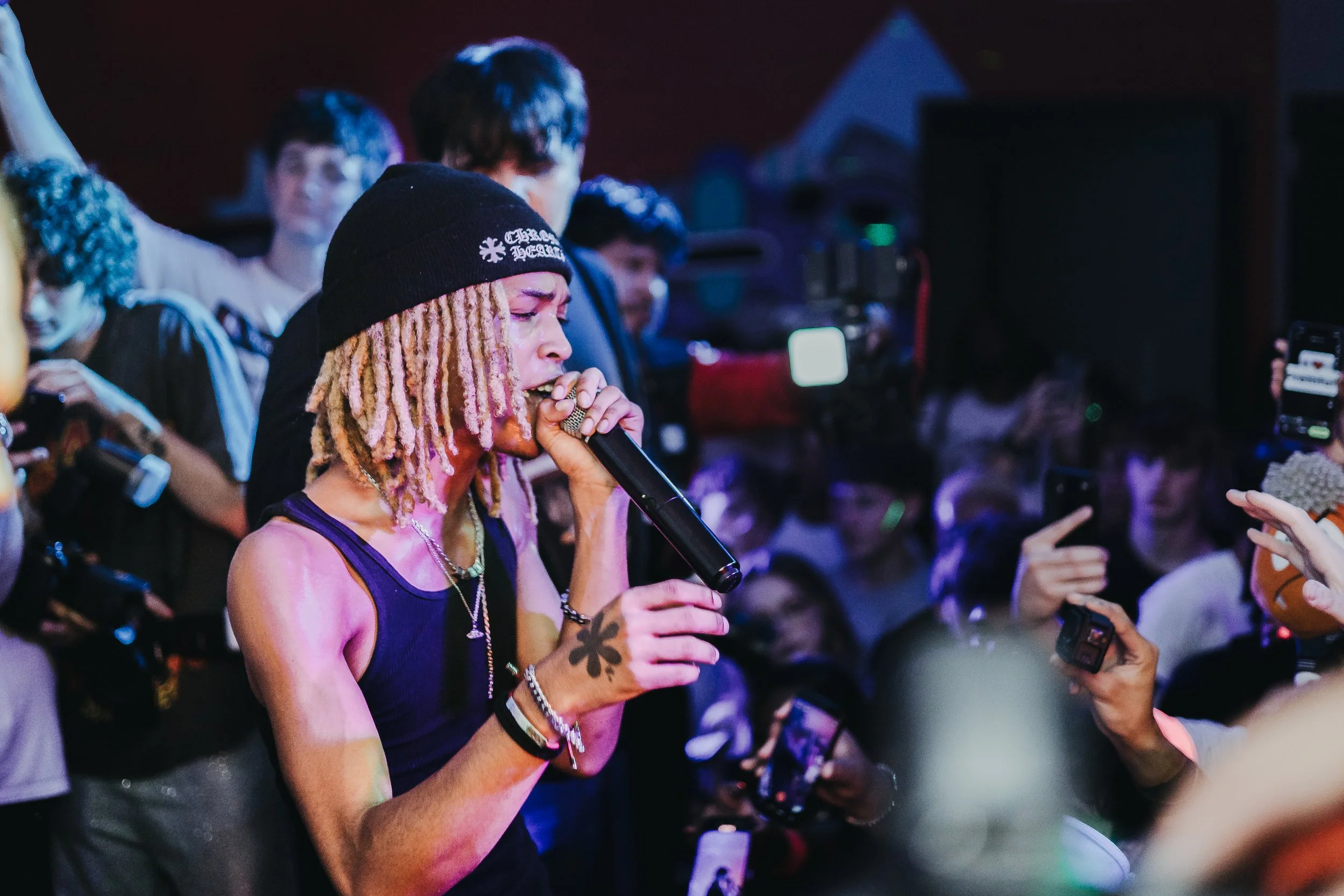 A performer with blonde dreadlocks, wearing a black beanie, singing into a microphone at a crowded event with people taking photos and videos.