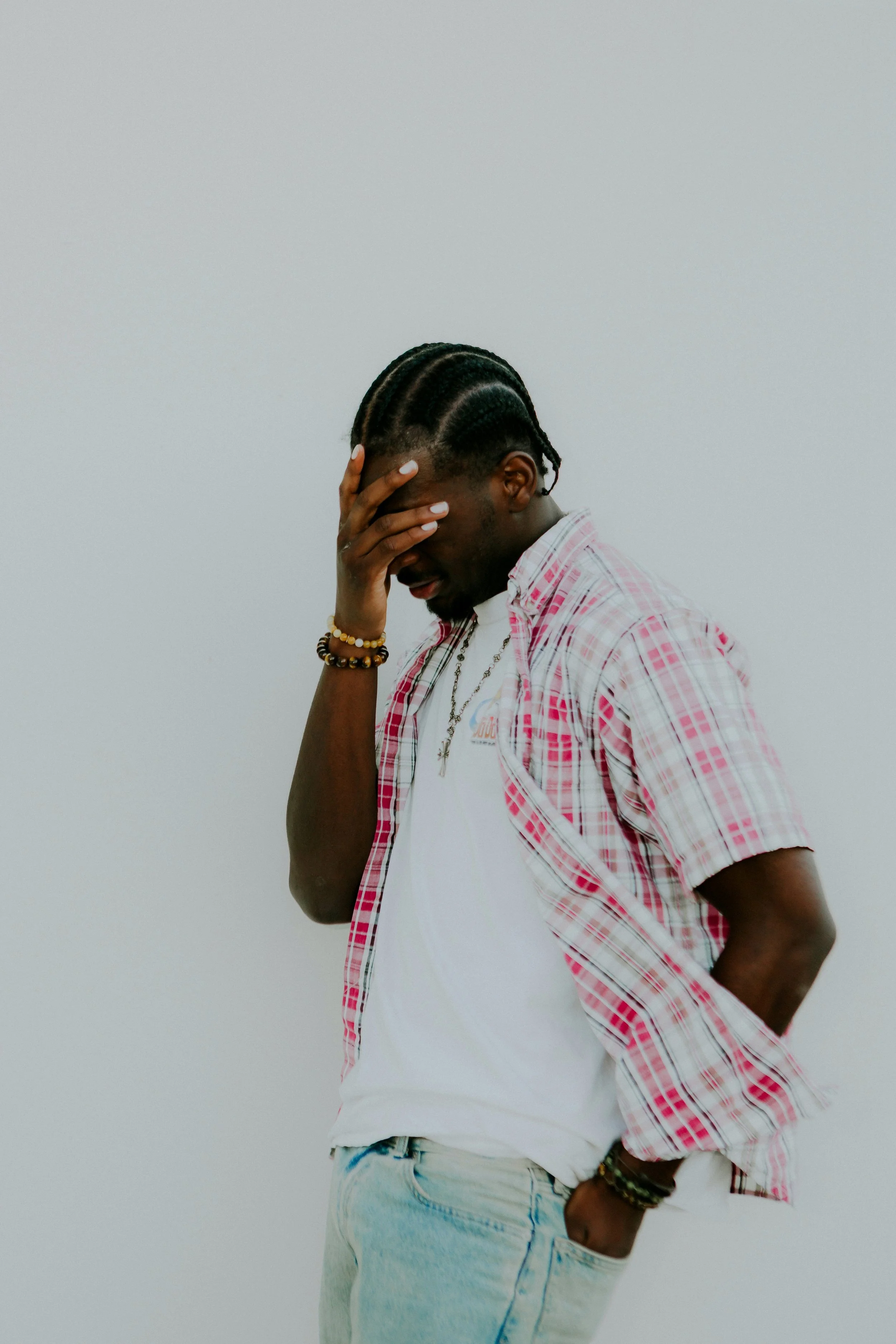 A man with braided hair, wearing a pink plaid shirt over a white t-shirt, stands against a plain background with his hand covering part of his face, looking downward.