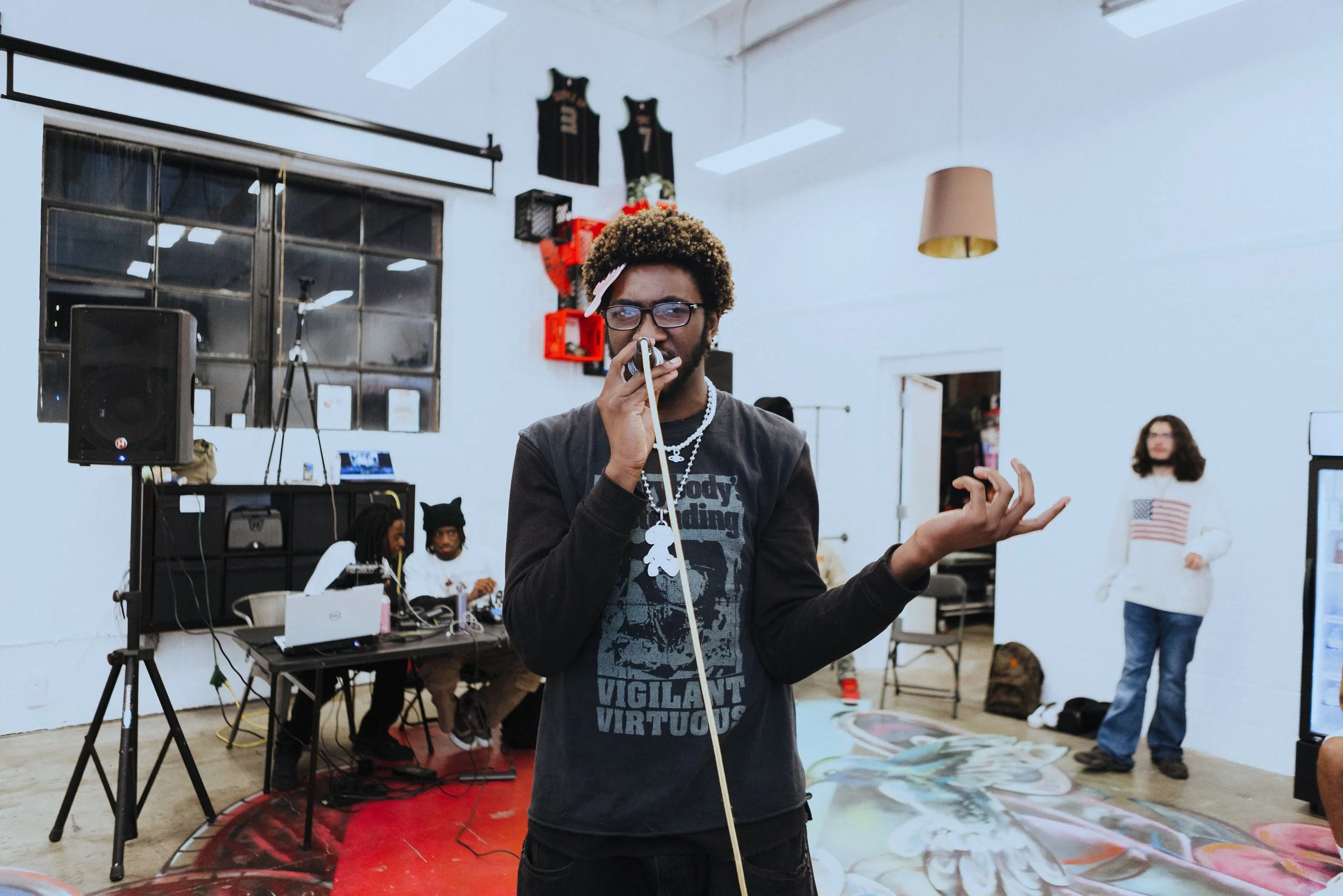 A young man with glasses and a curly hairstyle sings into a microphone, holding a strap in his left hand, in an indoor space with white walls and a red carpet. There are two people sitting at a table in the background with sound equipment, and a woman standing to the right in a white sweater with an American flag design.