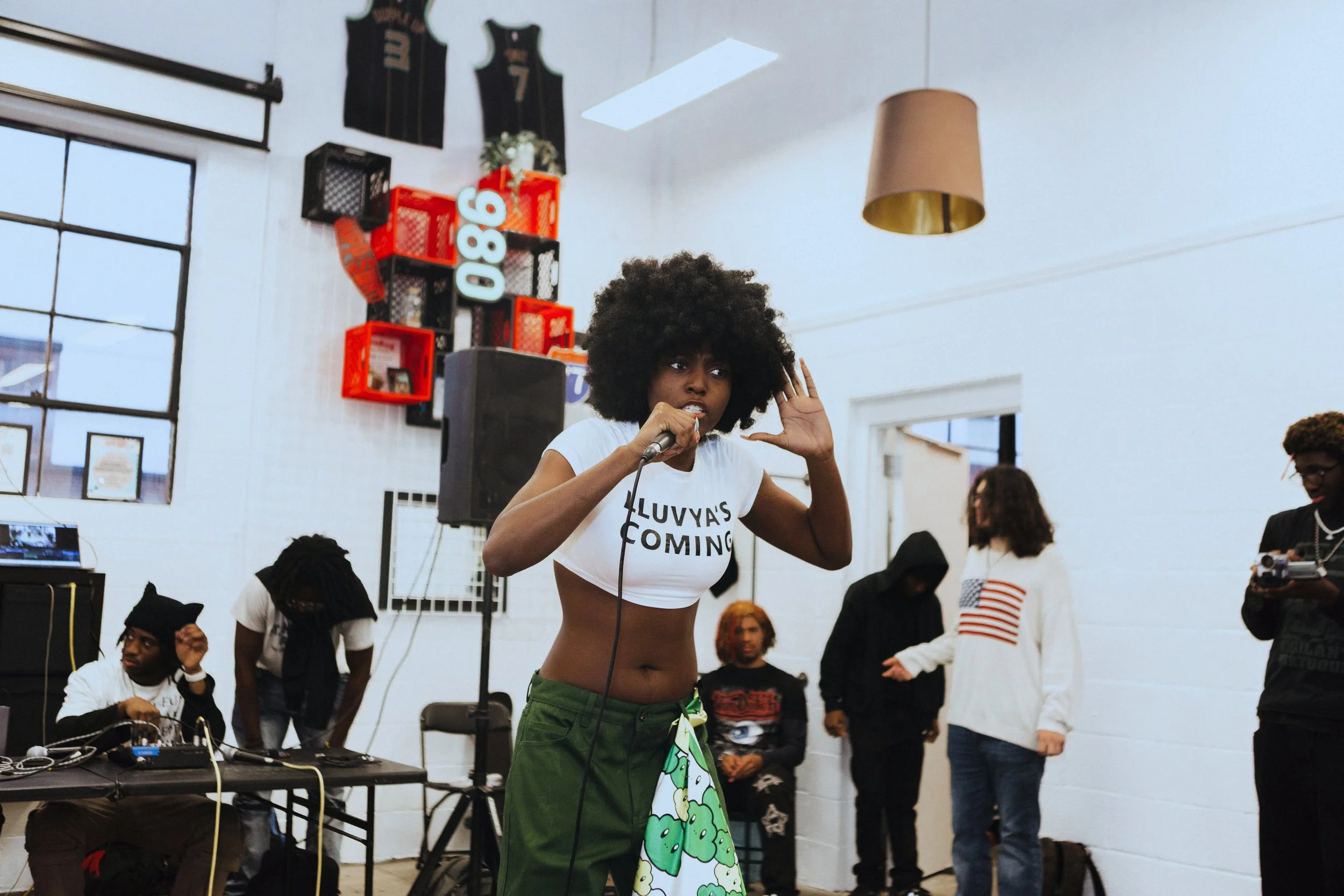 A woman with large curly hair speaking into a microphone at an indoor gathering, with people around her, some using phones or equipment.