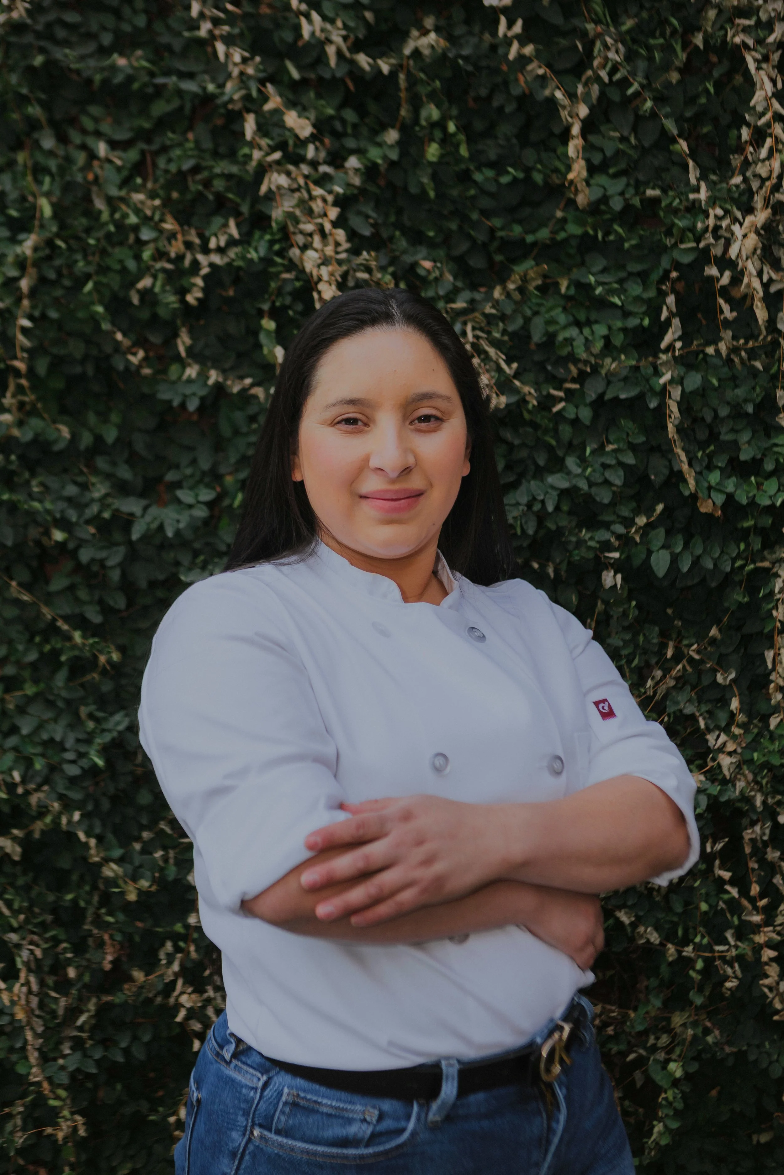 A woman in a white chef's coat standing outdoors in front of green leafy plants, with arms crossed and smiling.