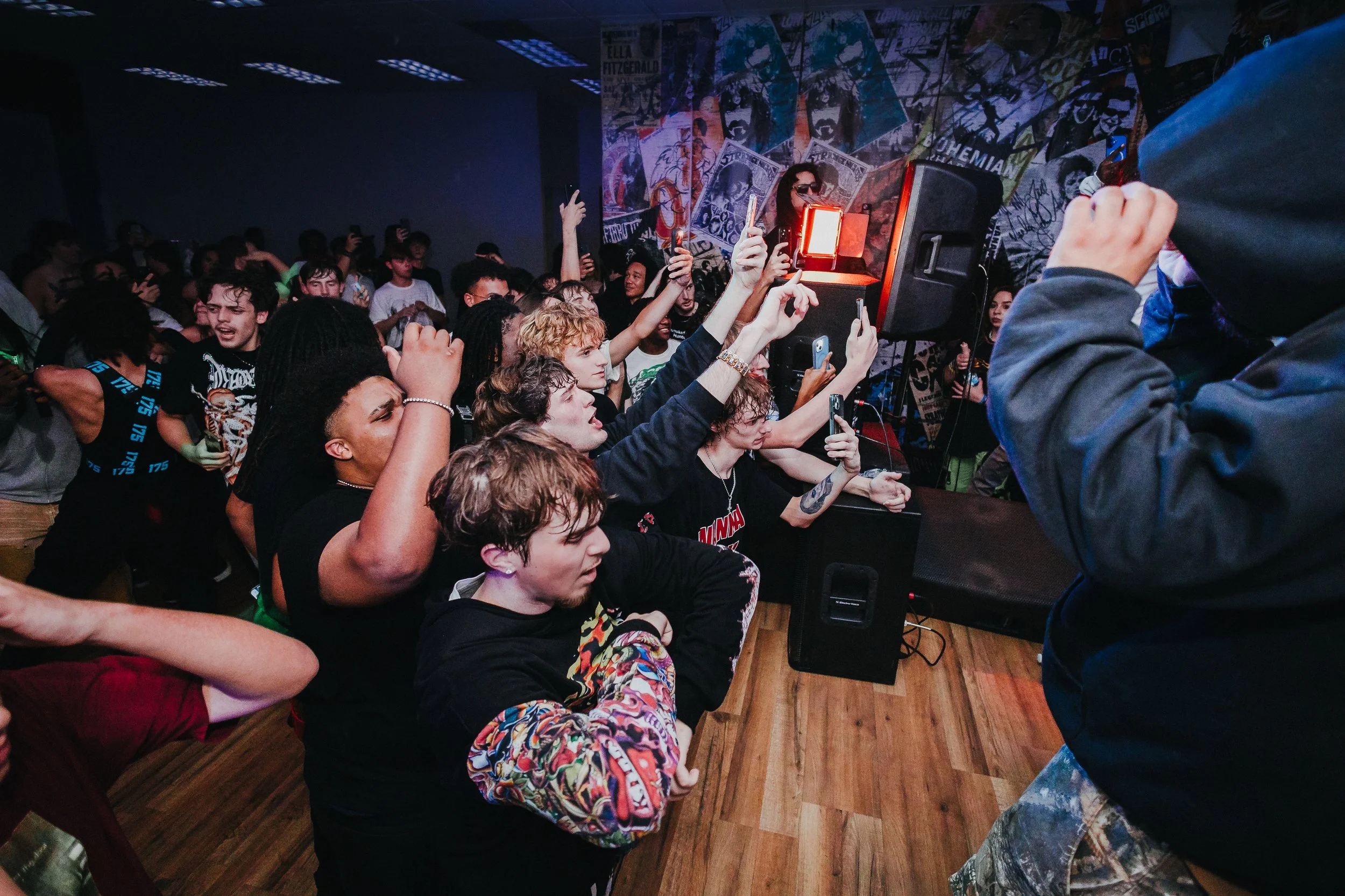 Crowd of young people at a live music concert, some taking photos or videos with smartphones, in a dimly lit indoor venue with graffiti-covered walls.