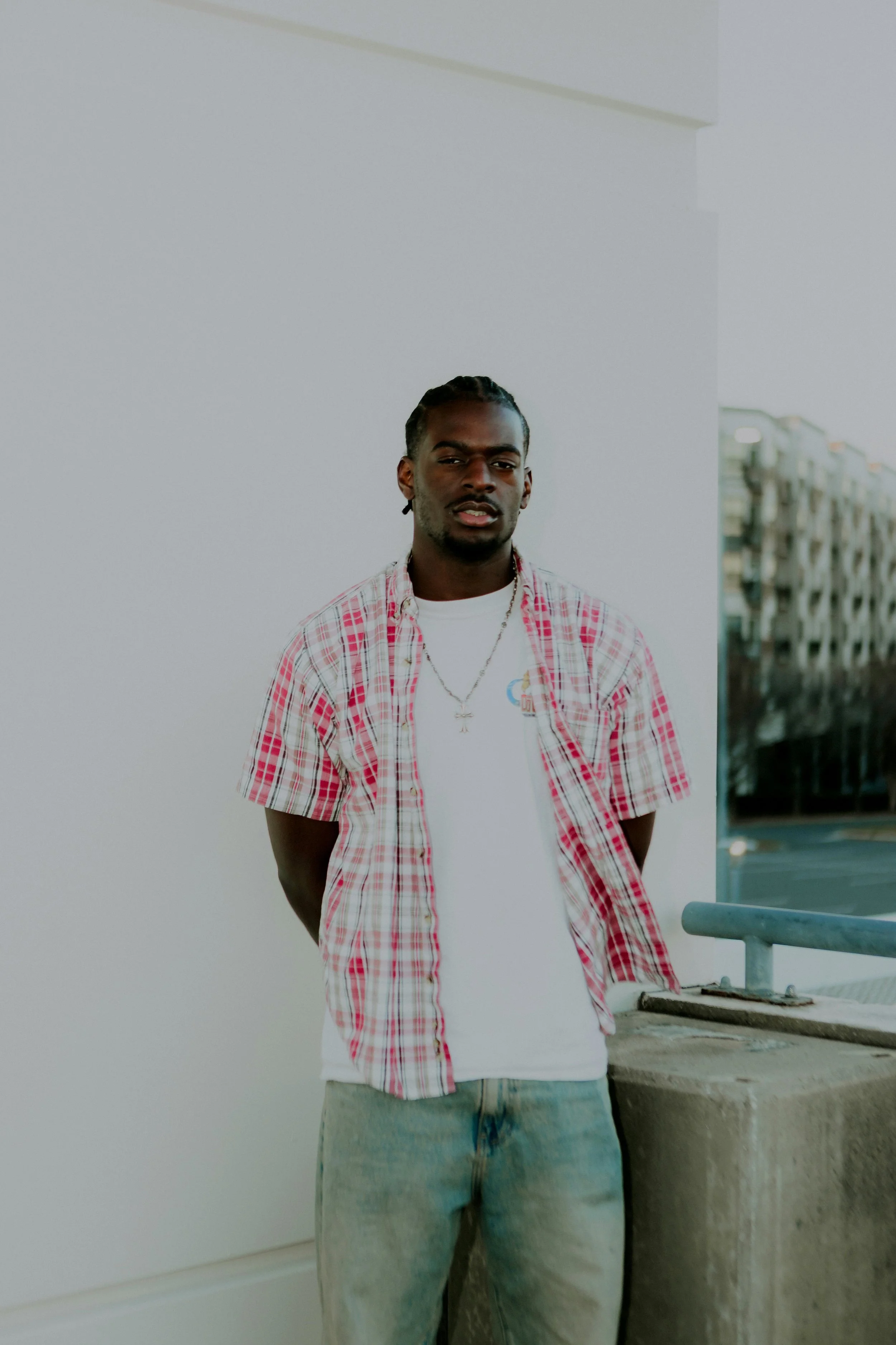 A young man with braided hair, wearing a white T-shirt, a red and white plaid shirt, and light blue jeans, standing outdoors near a concrete structure with a railing, with a background of buildings and a partly cloudy sky.