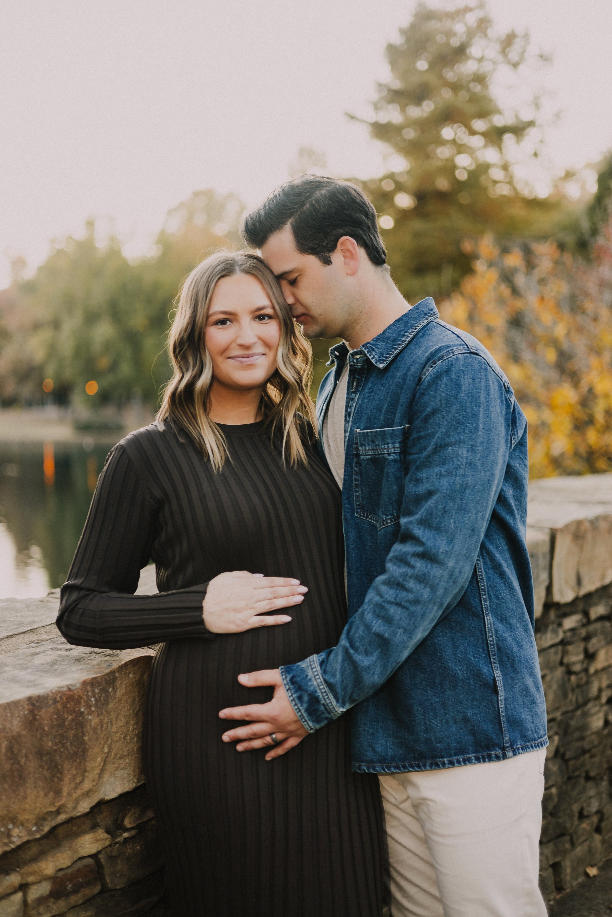 A couple outdoors during autumn, with the woman pregnant, standing by a stone wall near a river, with trees in fall foliage in the background.