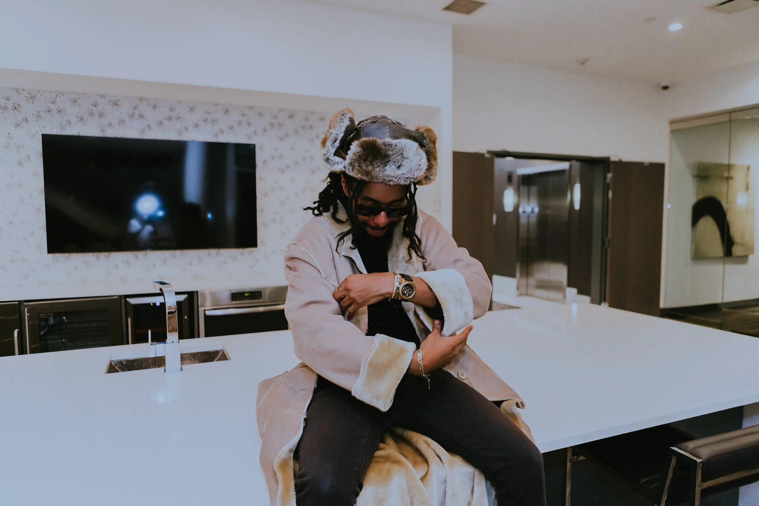 A man with dreadlocks and sunglasses wearing a tiger hat, a beige coat, and black pants, sitting on a countertop in a modern kitchen, looking down and smiling.