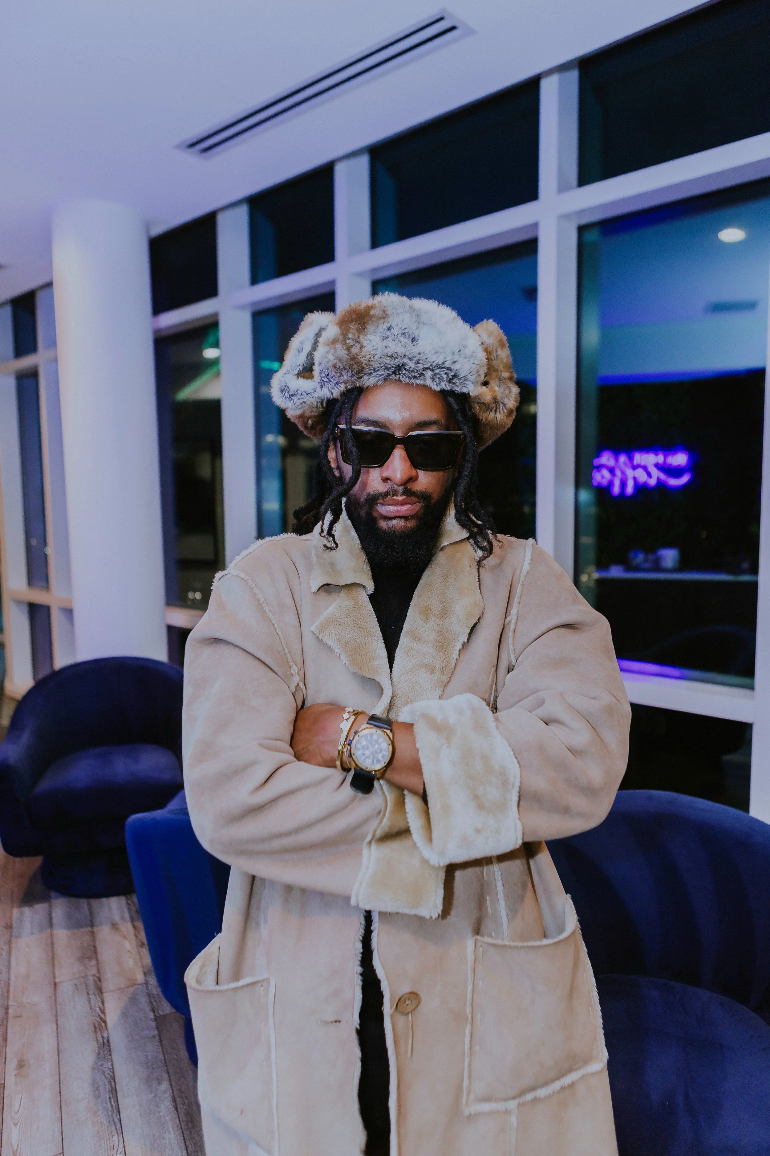 Man wearing a fur hat and sunglasses with crossed arms standing inside a modern building.