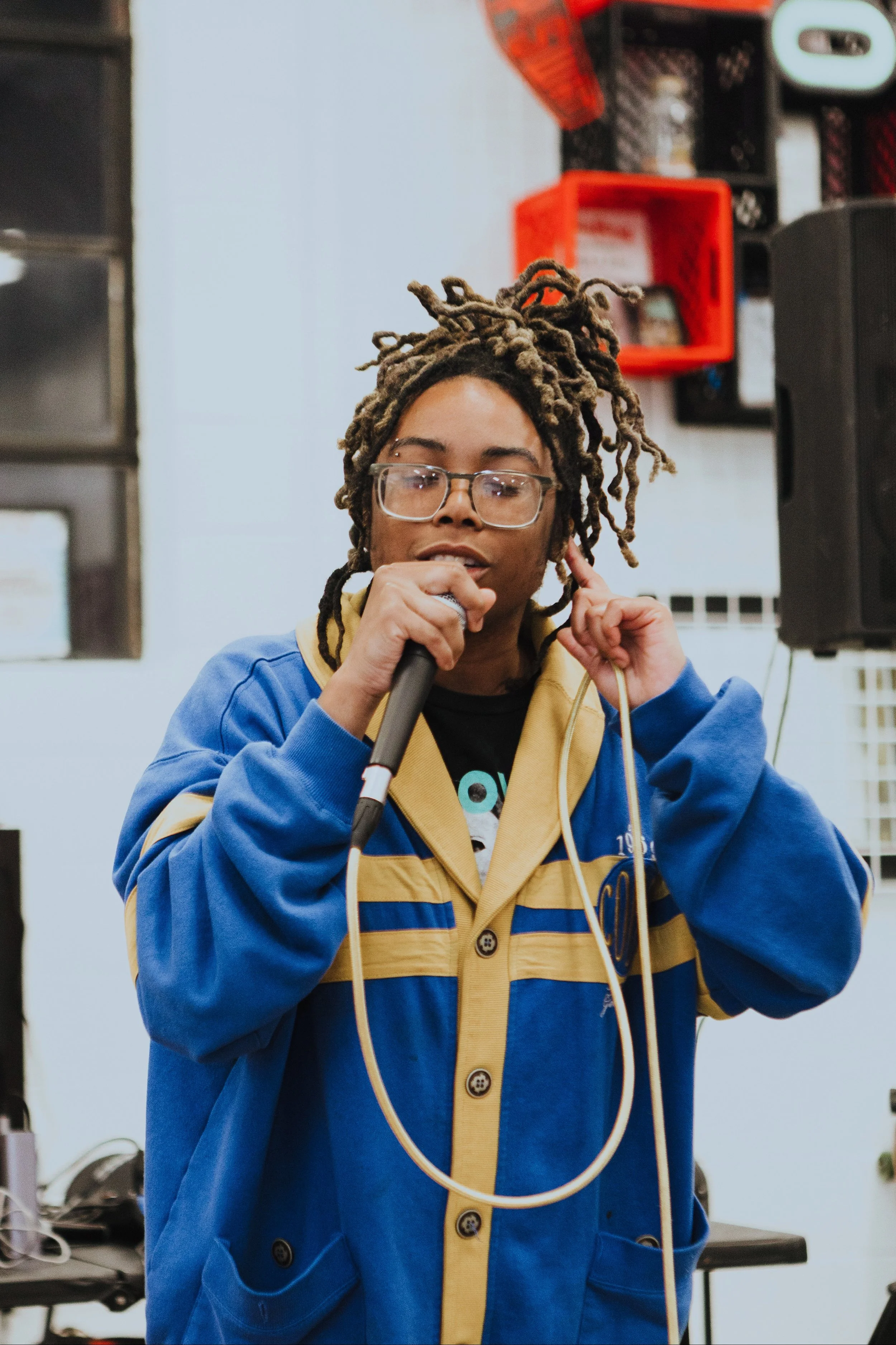 A person with dreadlocks and glasses holding a microphone, wearing a blue and yellow jacket, speaking or performing in a room with audio equipment and posters in the background.