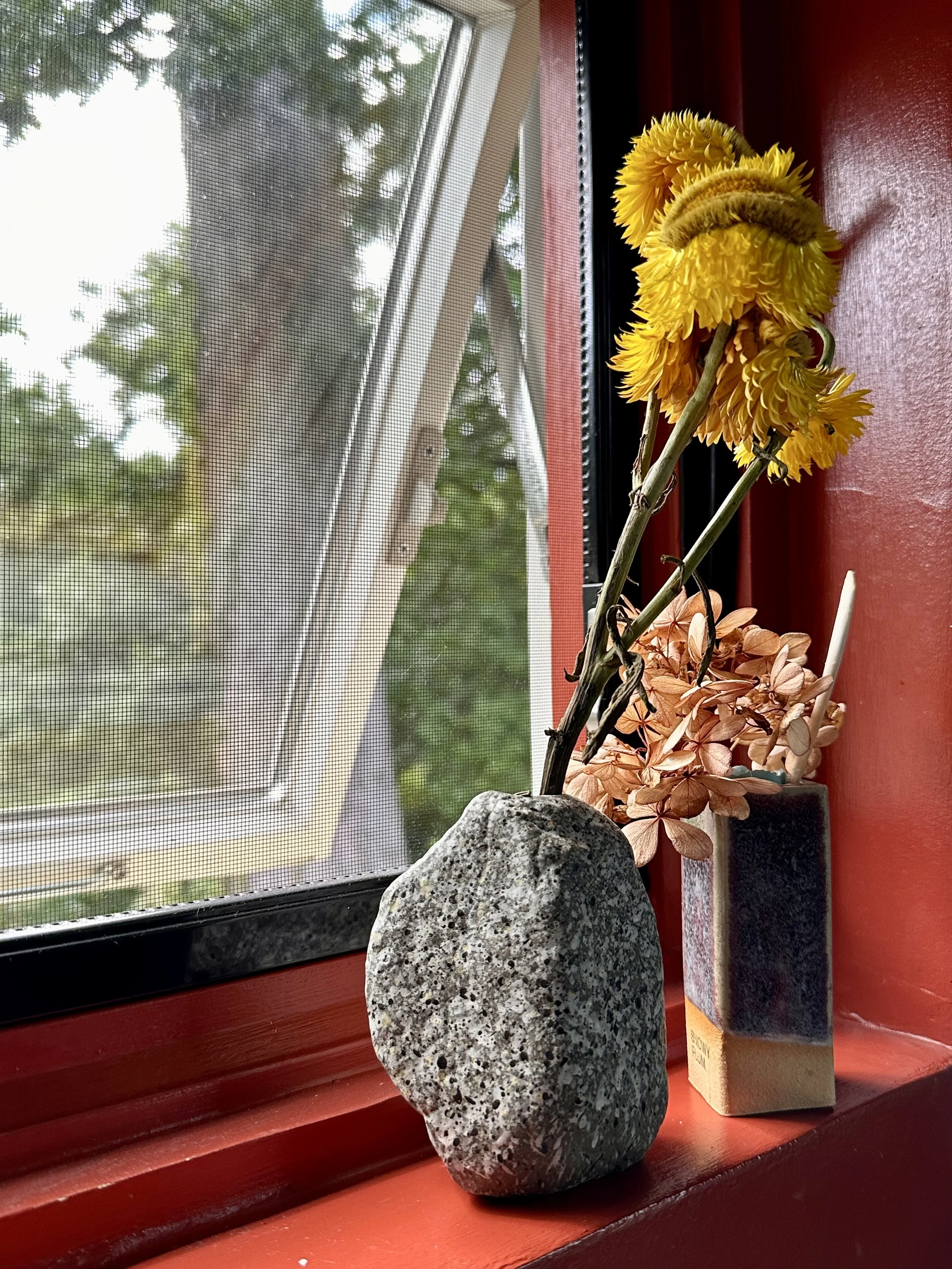A couple tiny vases holding a few soft blooms on a sunlit window ledge, backlit muted reds, yellows, and warm neutral tones