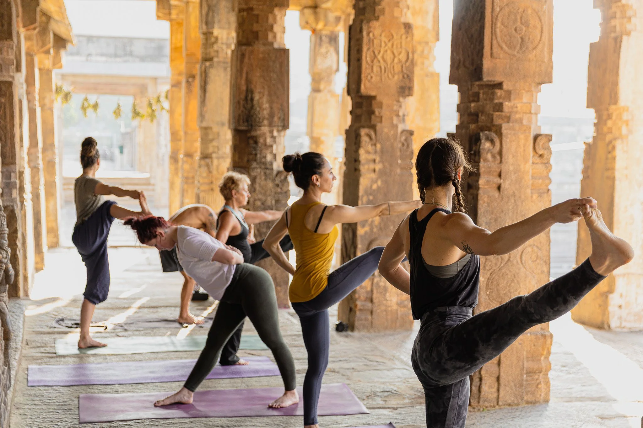 Group practicing standing yoga poses in a sunlit stone temple in Melkote, India