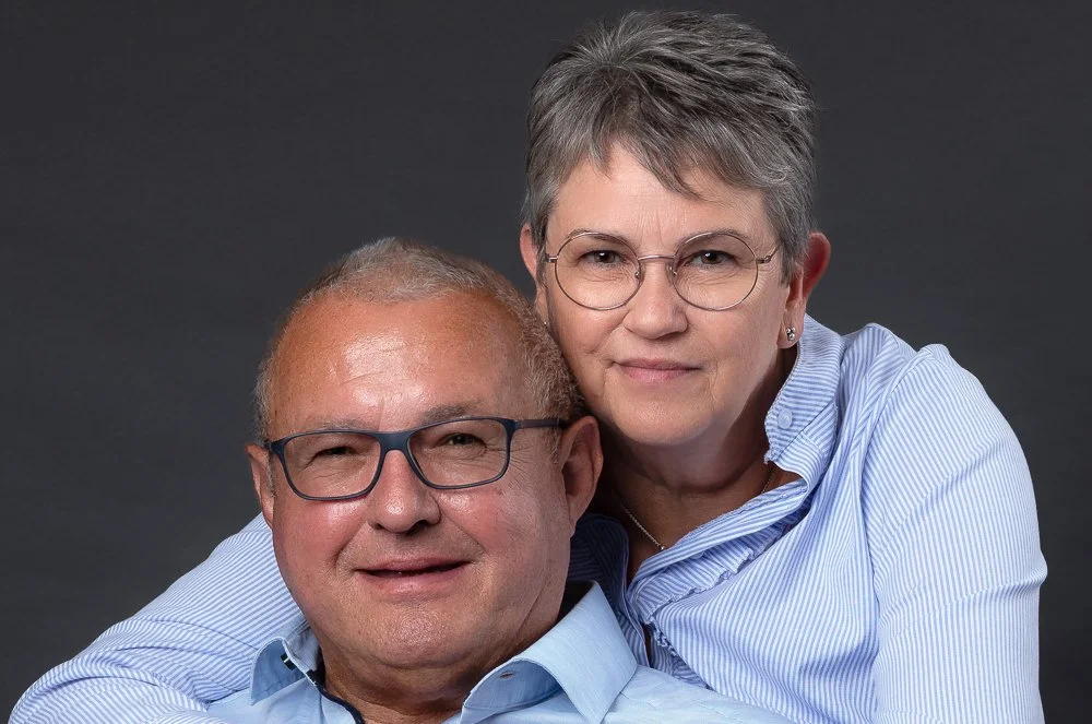 Et ældre par med briller, den mand har lyst hår og den kvinde har kort gråt hår, begge iført blåstribede skjorter, portræt mod en mørk baggrund.