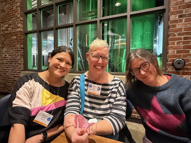 Three middle-aged women wearing conference badges and smiling inside a brick building.