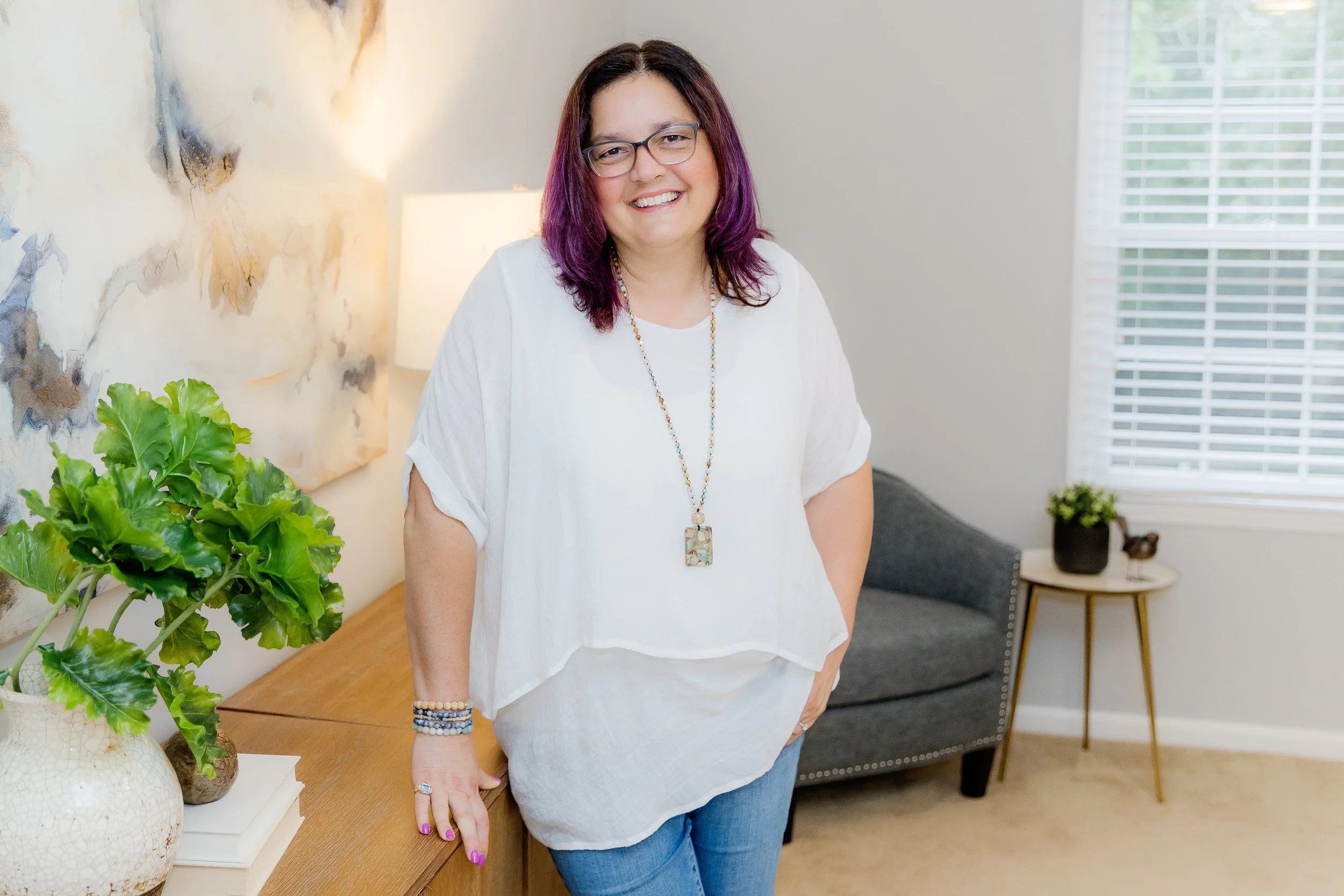 A woman with purple hair, glasses, and a white top sitting on a beige armchair in a bright living room with white walls and decorative shelves.