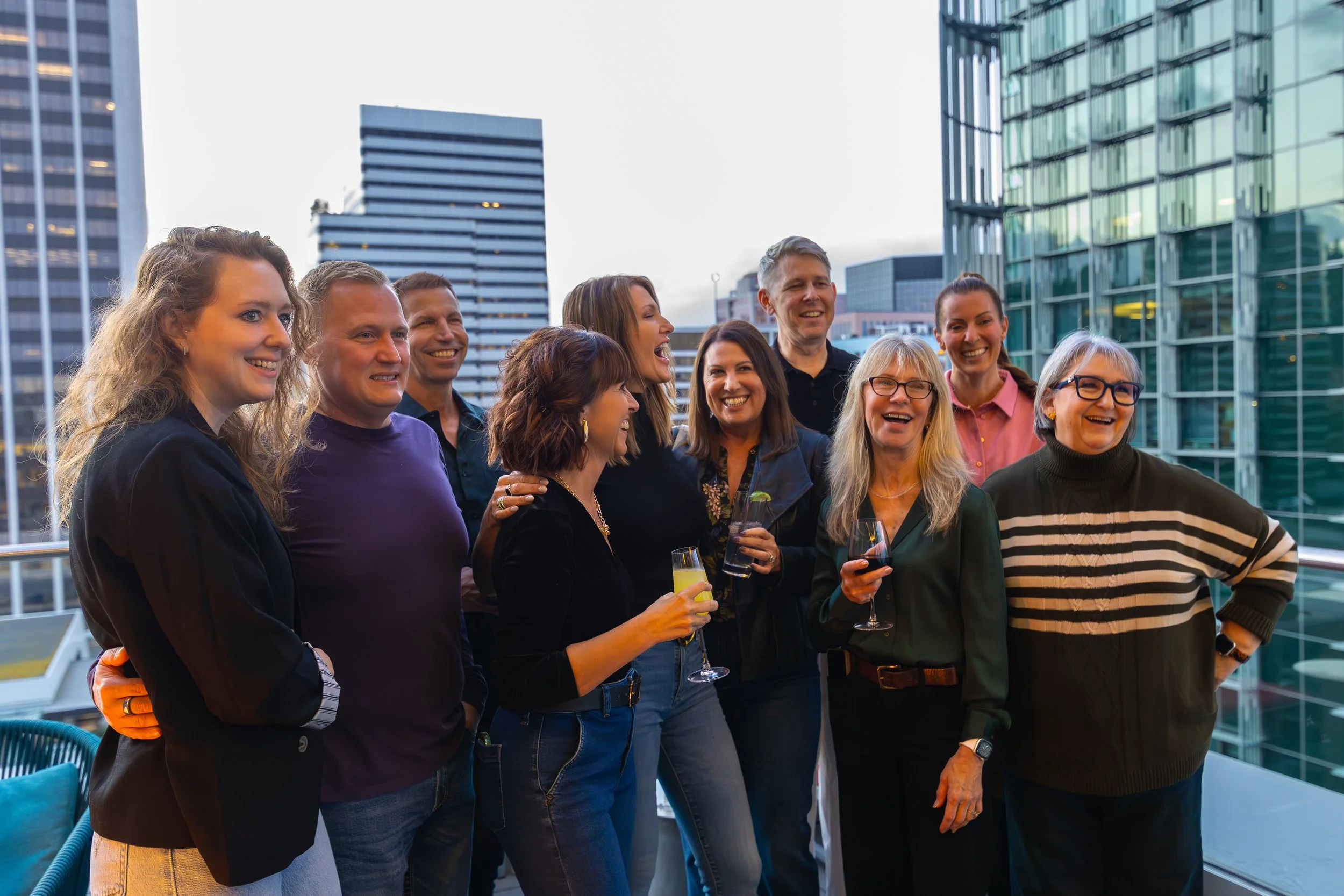 Group of people laughing and chatting at a rooftop party with city buildings in the background, some holding glasses of wine and drinks.