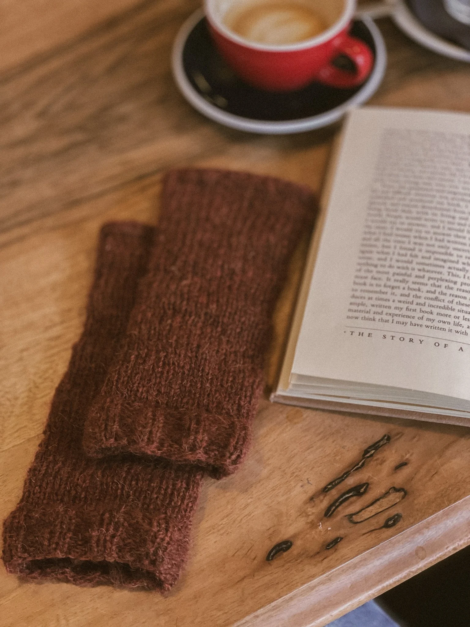 A wooden table with a pair of folded brown knit socks, an open book, and a red coffee cup on a black saucer.