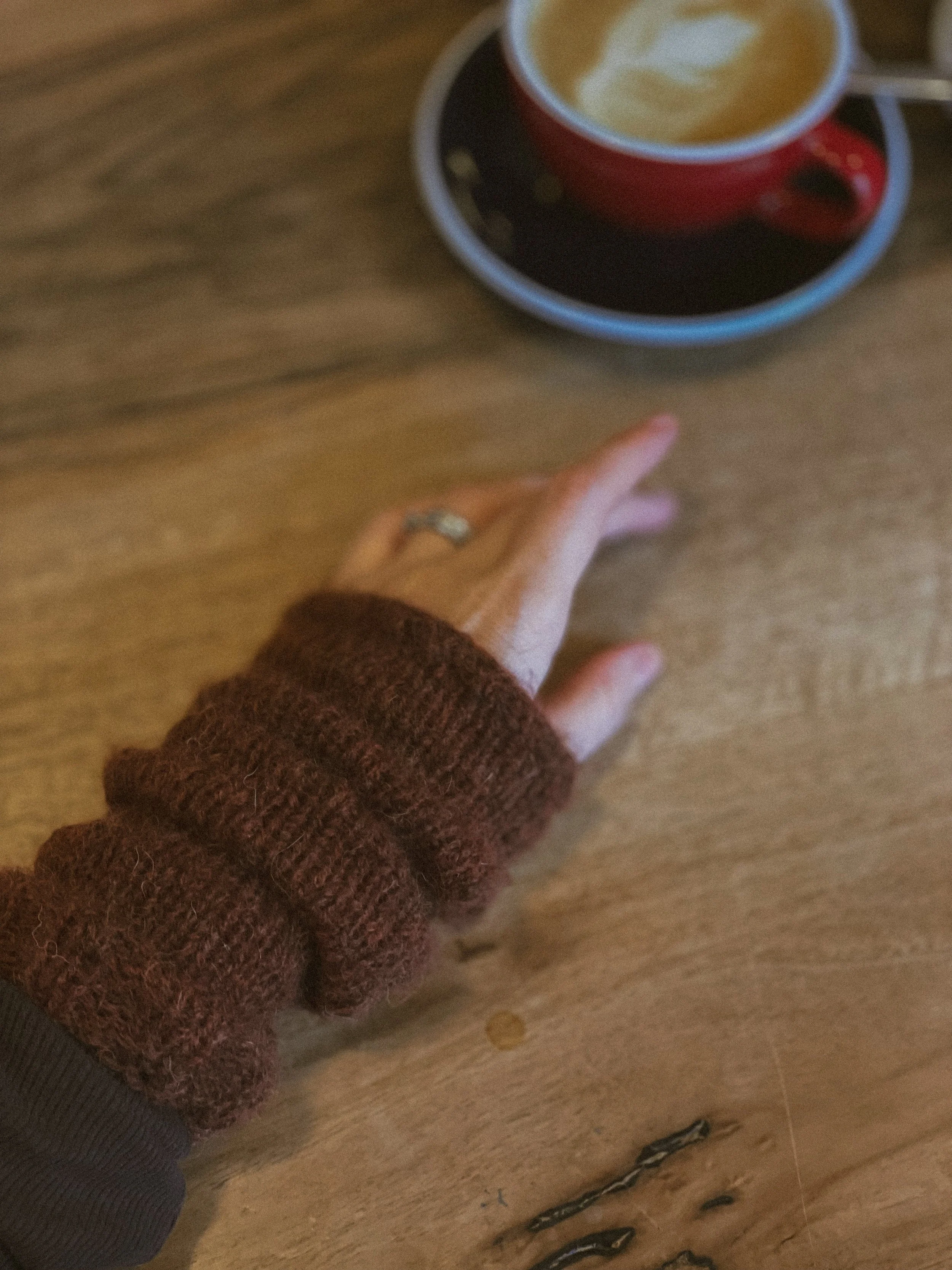 A person wearing a brown sweater reaching towards a red cup of latte with foam art on a wooden table.