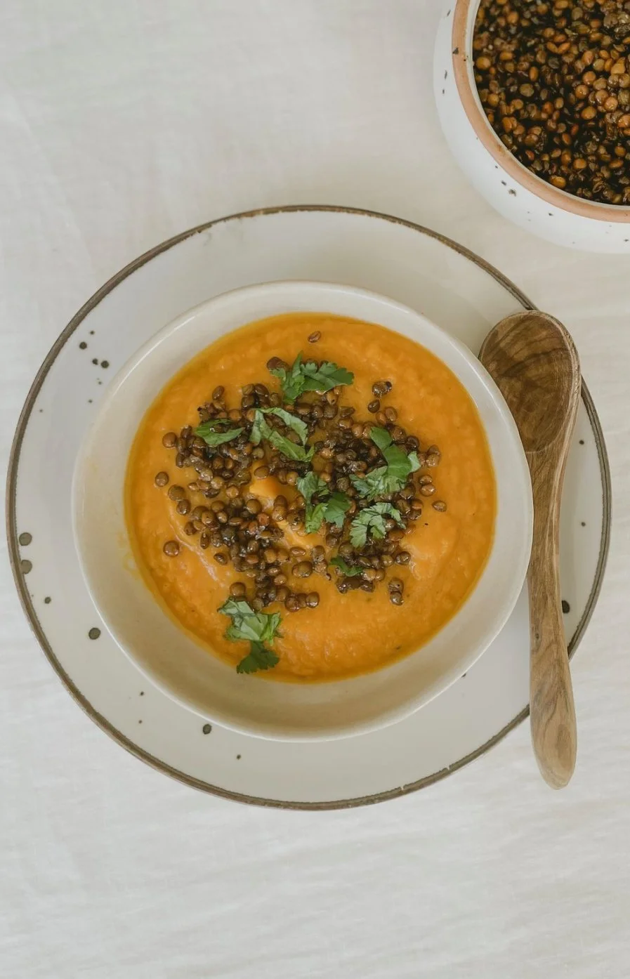 A bowl of pureed orange squash soup topped with roasted black lentils and chopped cilantro, served with a wooden spoon on a white plate, with a bowl of roasted lentils in the background.