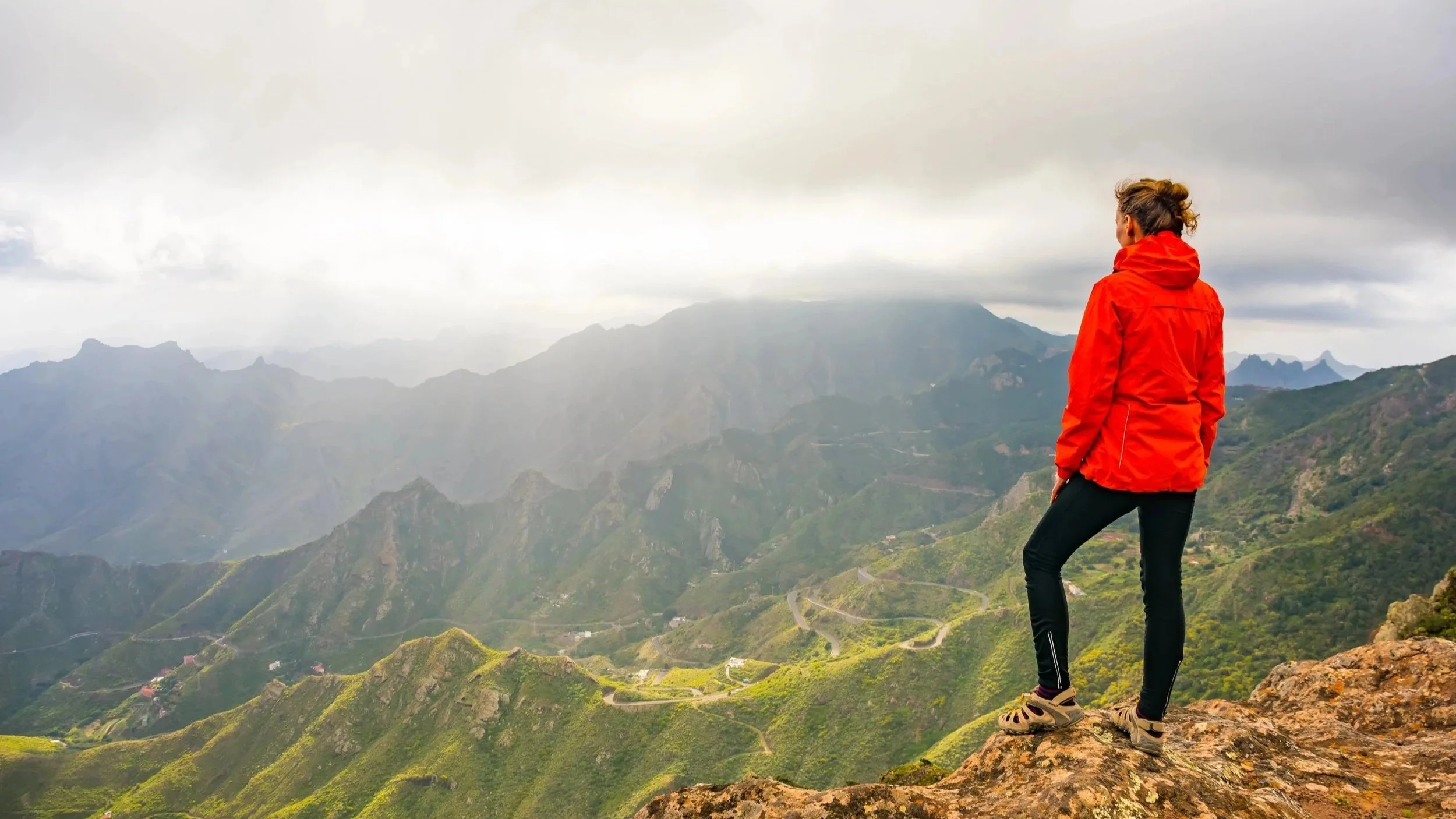 Woman trail blazer in a red parka and black pants standing at the edge of large canyon considering the possibilities