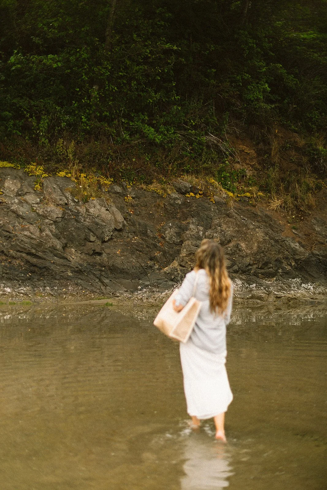 A woman walking in shallow water near a rocky shoreline with green foliage on a hillside in the background.