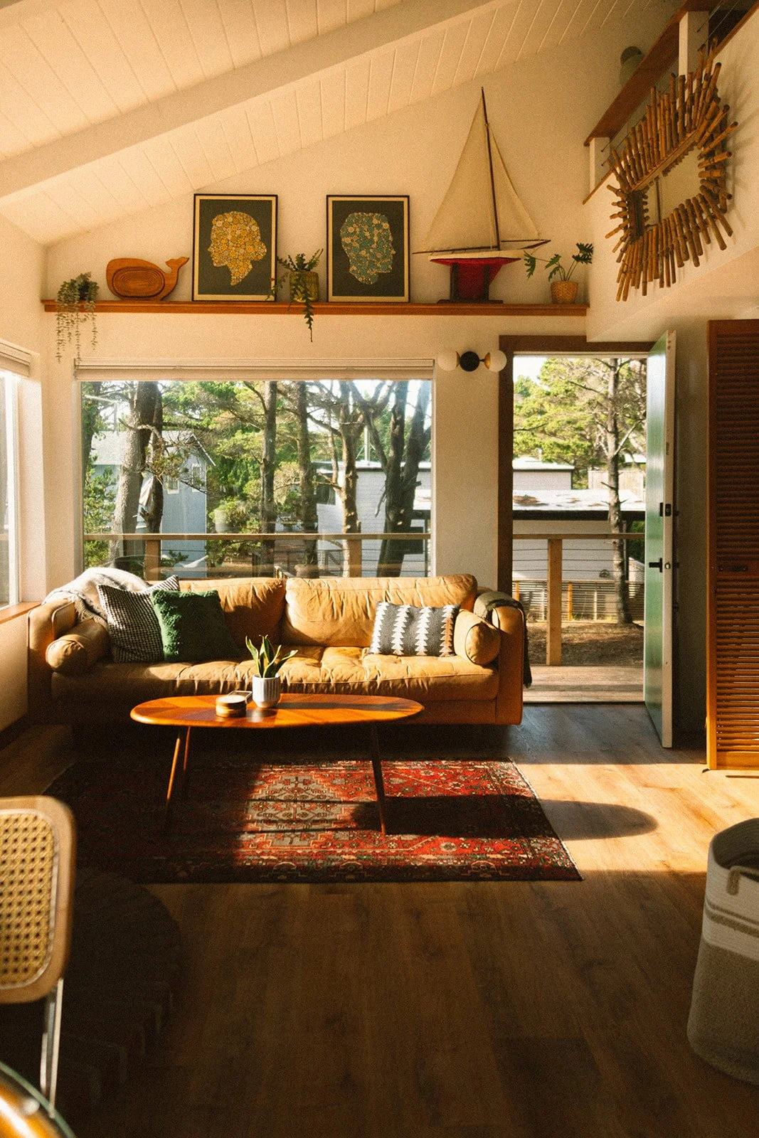 Sunlit living room with a tan sofa with green and patterned cushions, a wooden coffee table with a plant, large windows, and a door opening to a small deck with trees outside. Decorations include artworks, a model sailboat, and a decorative mirror.
