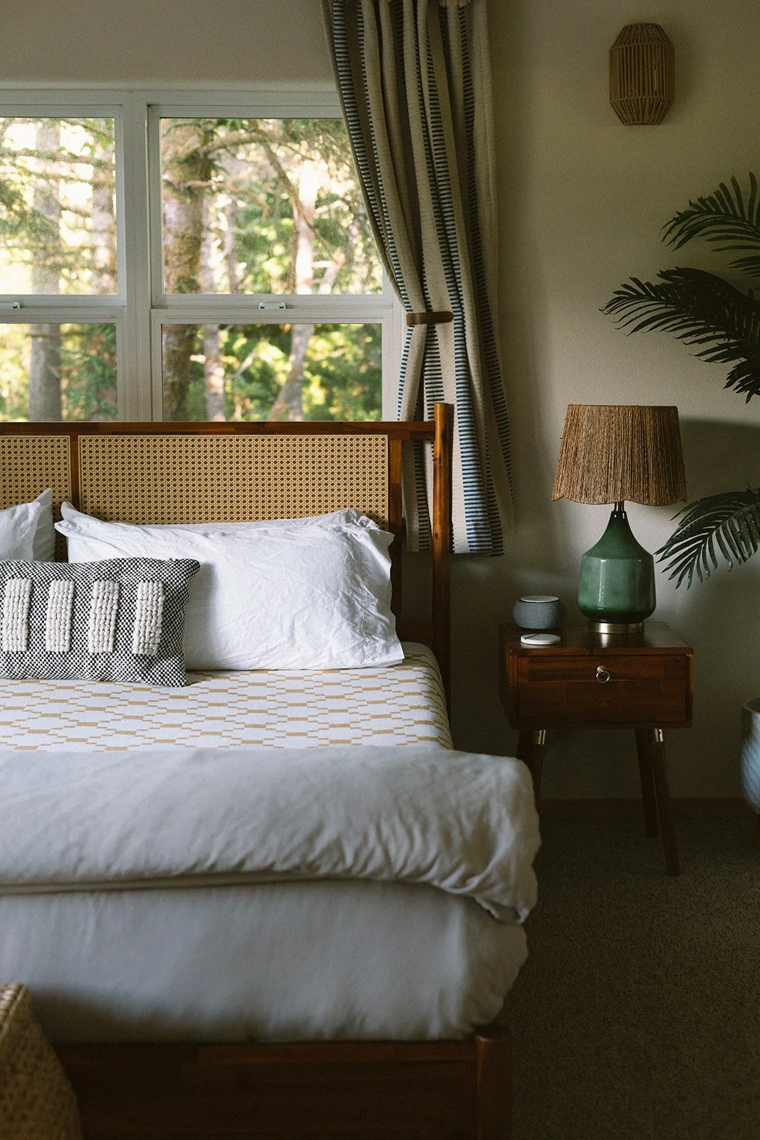 A cozy bedroom with a wooden bed frame, white bedding, and decorative pillows. Large window with striped curtains overlooks a wooded outdoor landscape. A bedside table holds a green ceramic lamp, a small gray speaker, and a remote. There's a potted p