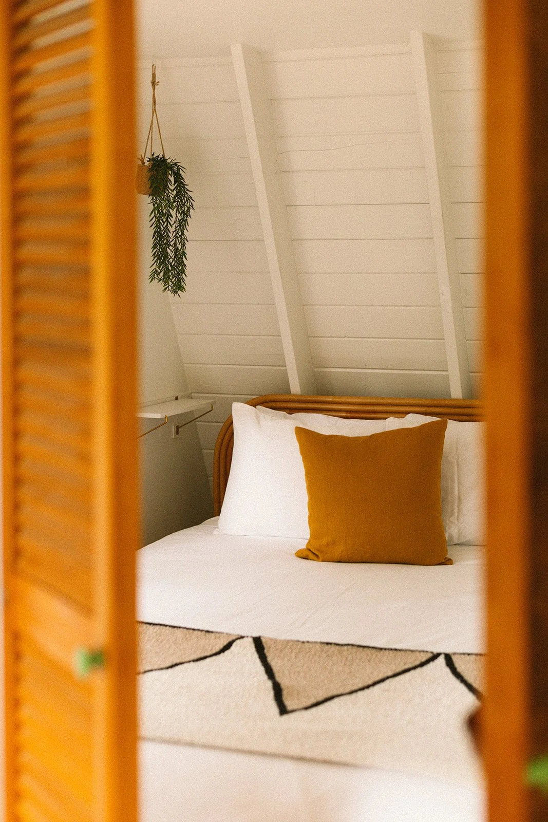 A cozy bedroom with a bed that has a white sheet and a mustard-colored pillow, seen through a wooden louvered door. The ceiling is angled with white wooden paneling, and there is a small hanging plant in the corner.