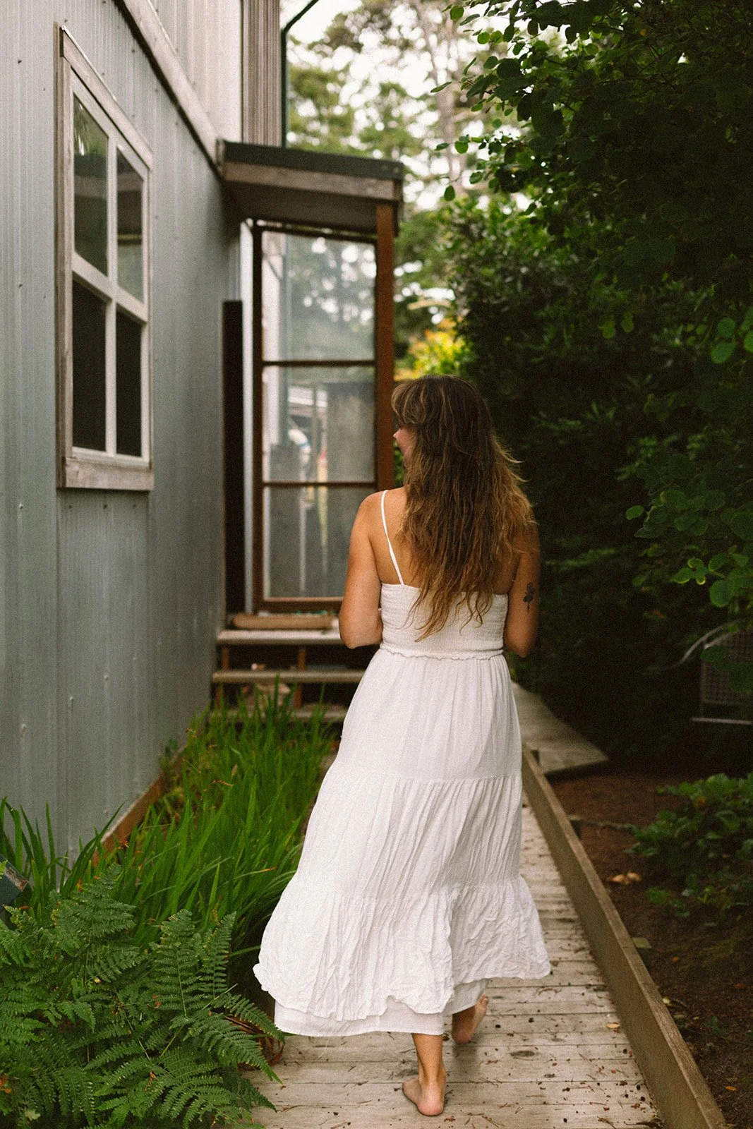 A woman with long wavy hair in a white sundress walking barefoot on a wooden pathway next to a house with gray siding, surrounded by green plants and trees.