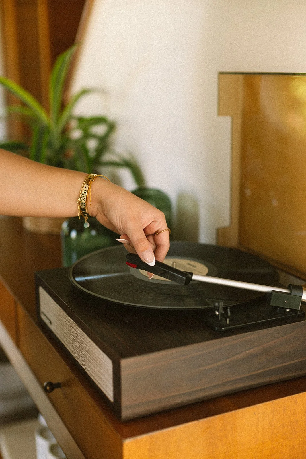 A person with a gold bracelet and ring on their hand adjusting the tone arm on a vintage turntable playing a vinyl record.